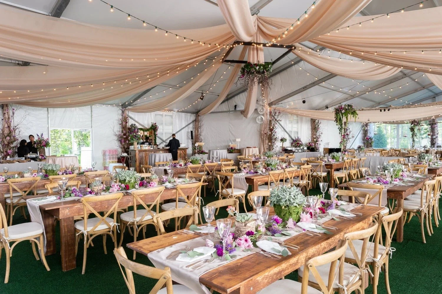 Wedding reception inside a large tent with draped fabric and string lights, decorated with floral arrangements on tables and hanging from the ceiling, set up for guests with wooden tables, chairs, and glassware.
