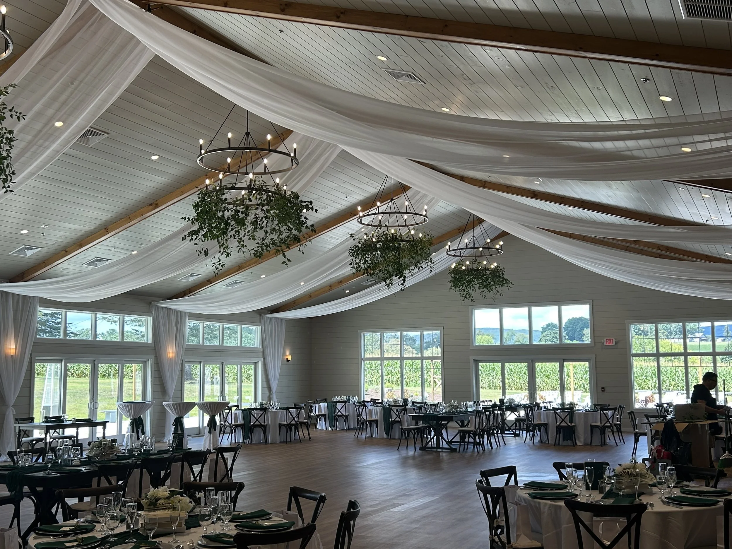 Large spacious banquet hall with high wood ceiling, white drapes, and large windows showing greenery outside. Round tables are set with white tablecloths, glassware, and centerpieces, and black chairs surround each table. Chandeliers and hanging greenery decor adorn the ceiling.
