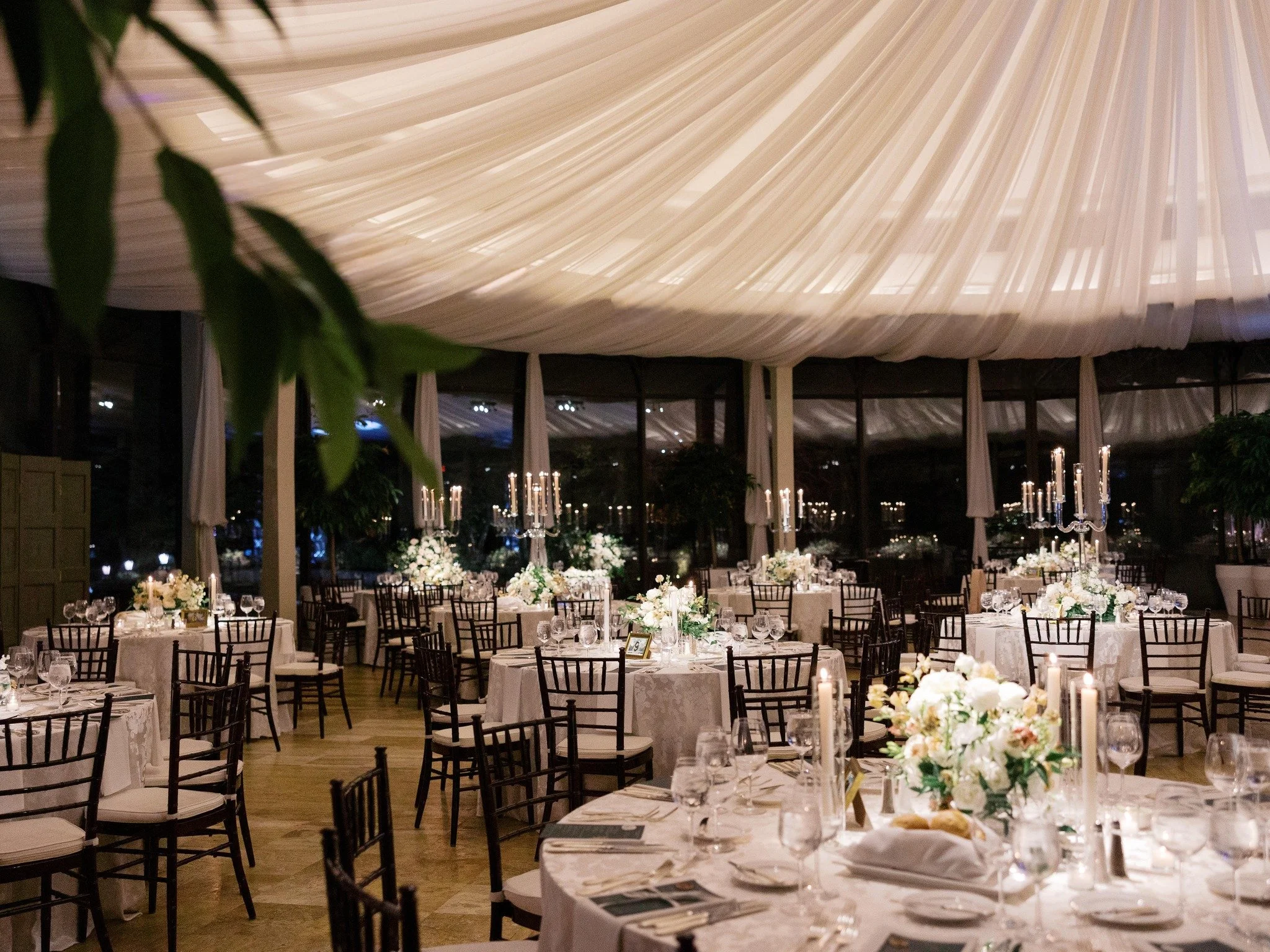 Elegant wedding reception inside a tent with round tables decorated with white tablecloths, floral centerpieces, glassware, and candles, ceiling draped with white fabric.