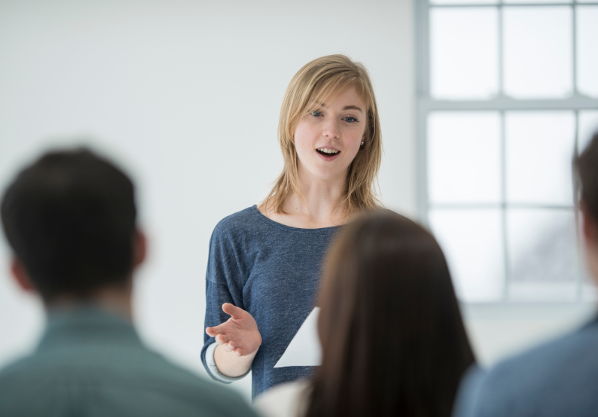 A young woman giving a presentation or speech to a group of people in a bright room with large windows.