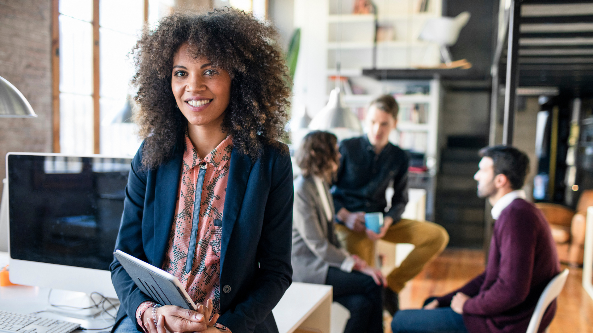 A smiling woman with curly hair holding a tablet in a modern office setting, with three colleagues in the background engaged in conversation.