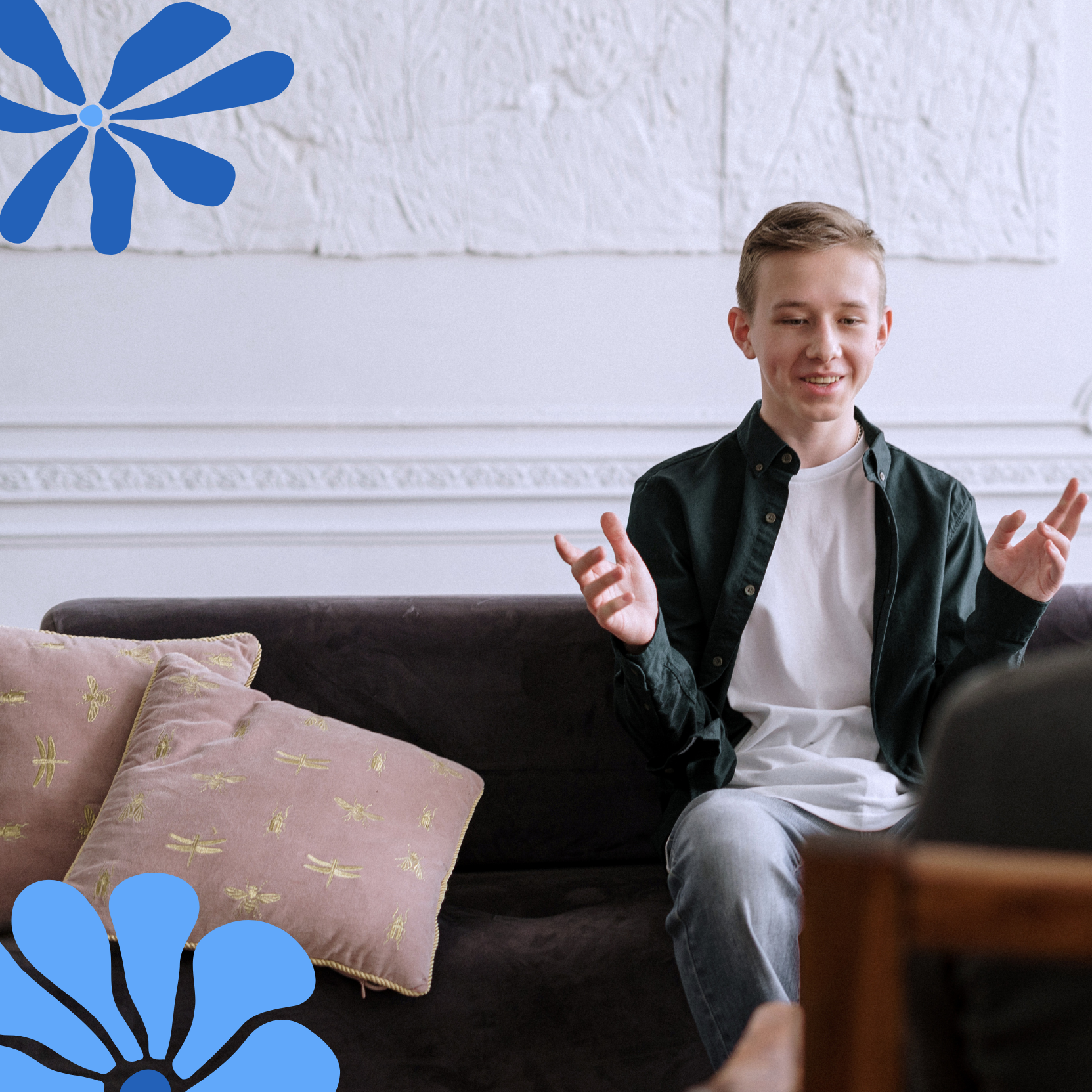 A teenage boy sitting on a couch, smiling and gesturing with his hands, in a room with white textured walls and pink pillows with gold embroidery on the couch. Blue floral designs are in the corner of the image.