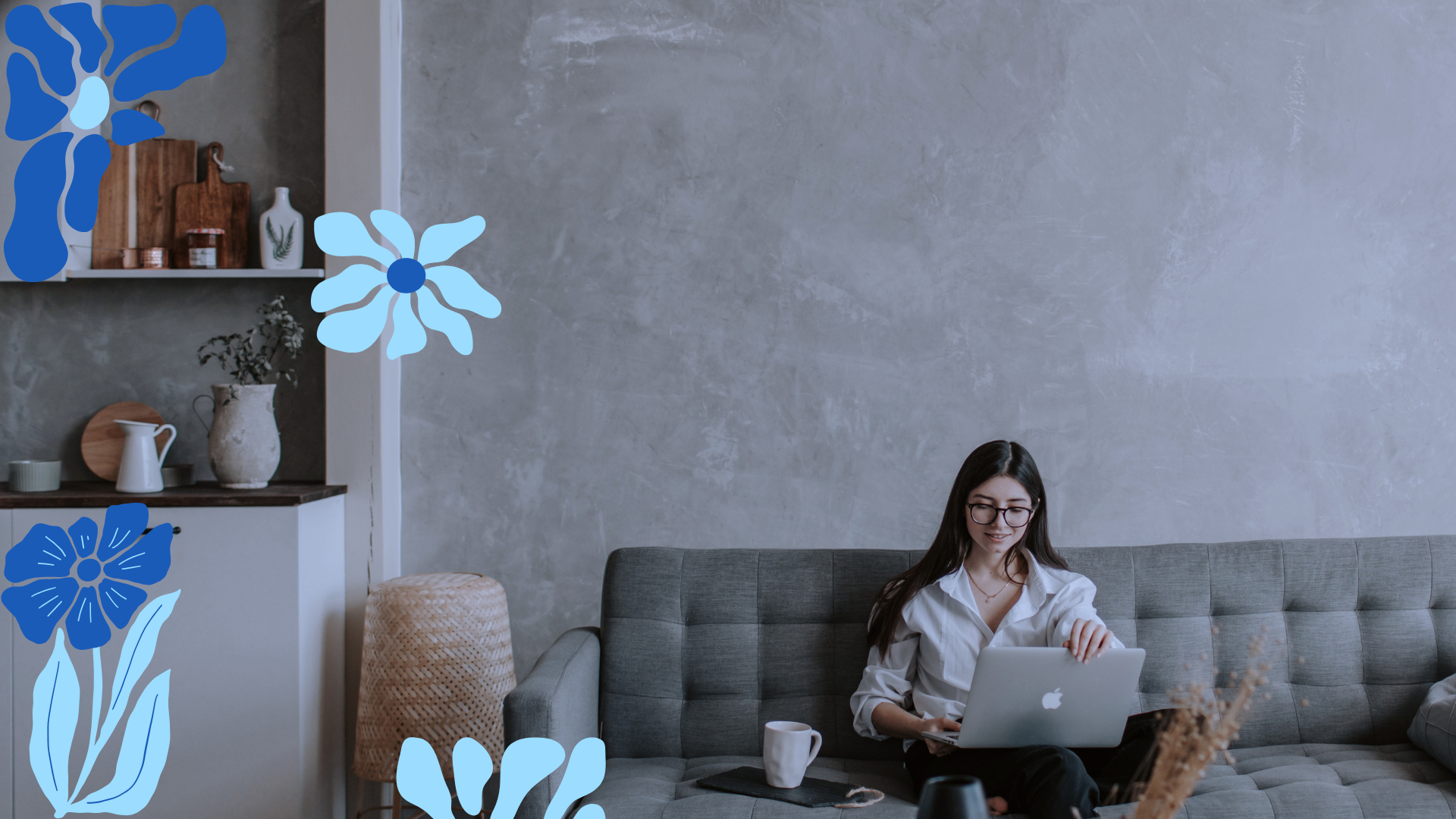 A woman with long dark hair and glasses sitting on a gray couch, working on a laptop. There is a white mug and a notebook on the table in front of her. The room has a minimalist decor with a gray wall, a white shelf with decorative items, and a woven stool.