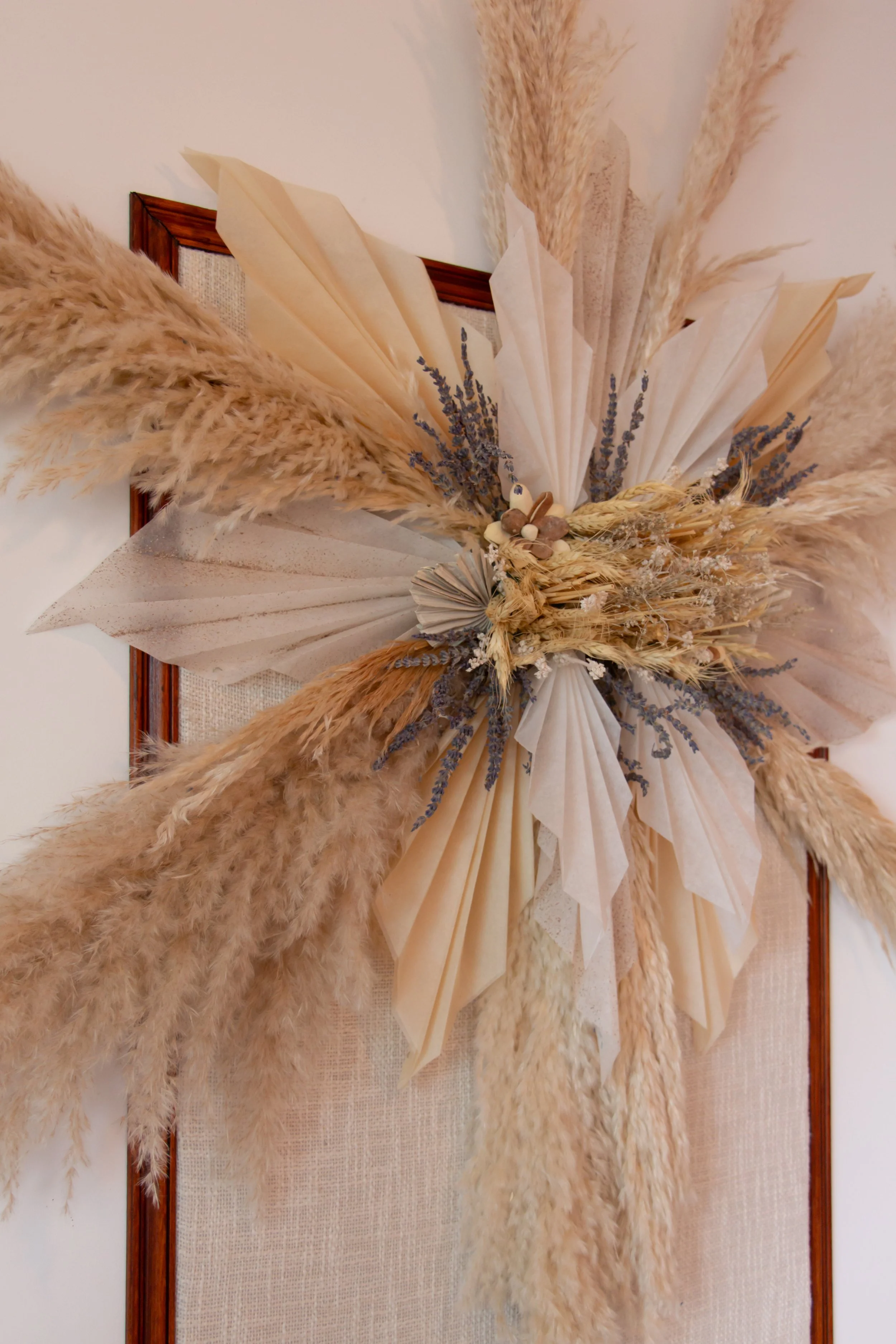 A decorative wall arrangement with dried pampas grass, lavender, and woven elements, displayed on a wooden frame with fabric background.