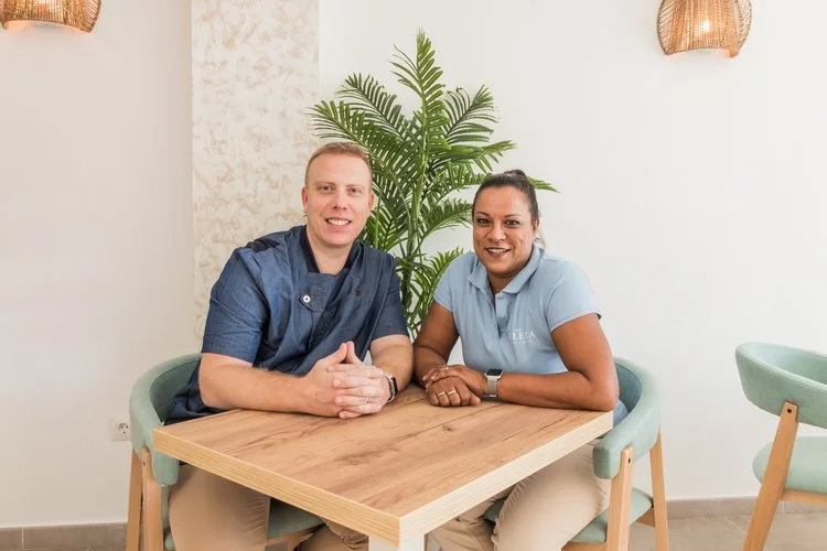 Two people sitting at a wooden table in a room with white walls and a large green plant, smiling at the camera.
