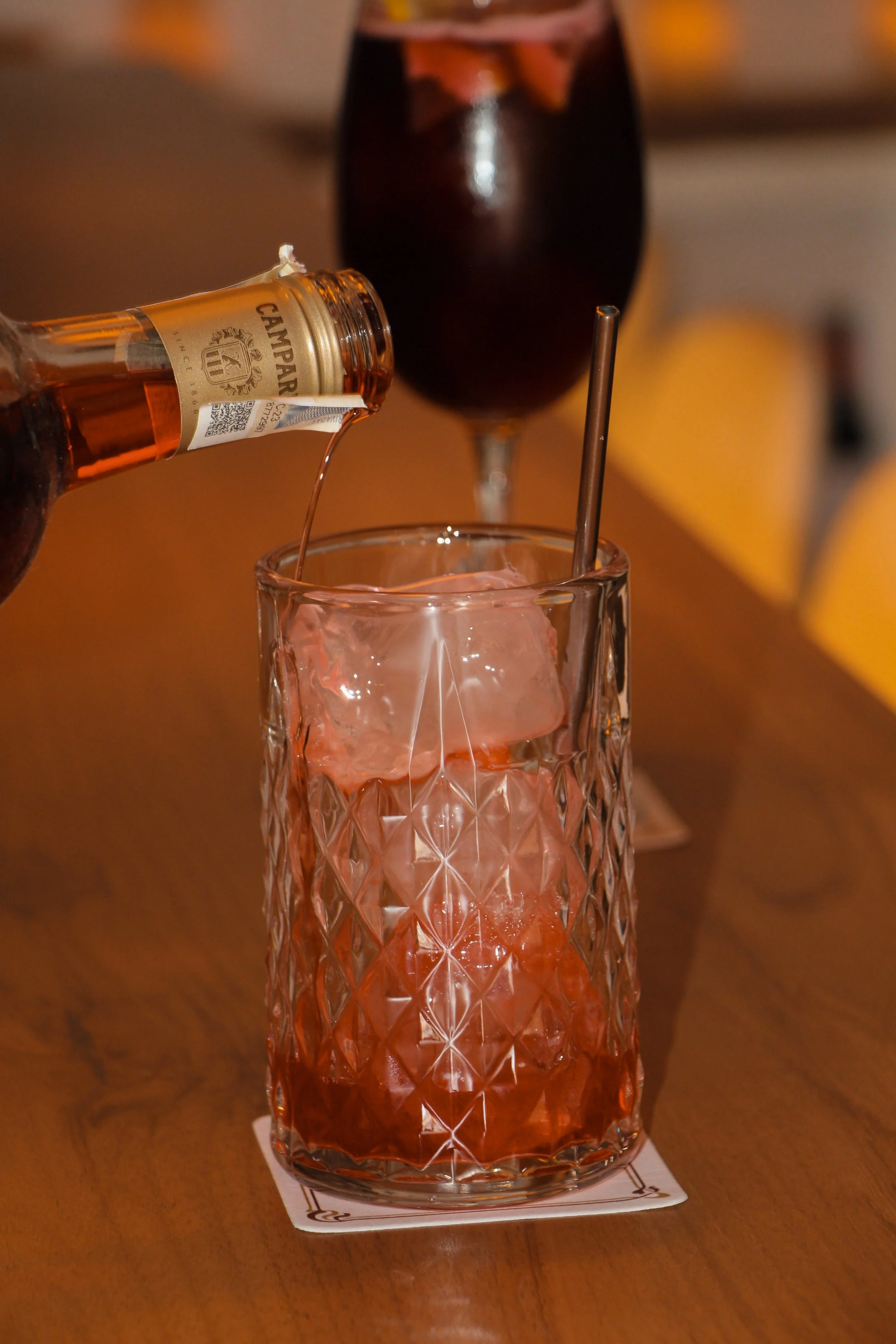 A glass of red cocktail with ice cubes, being poured from a small bottle, with a dark beverage in a wine glass in the background, on a wooden table.