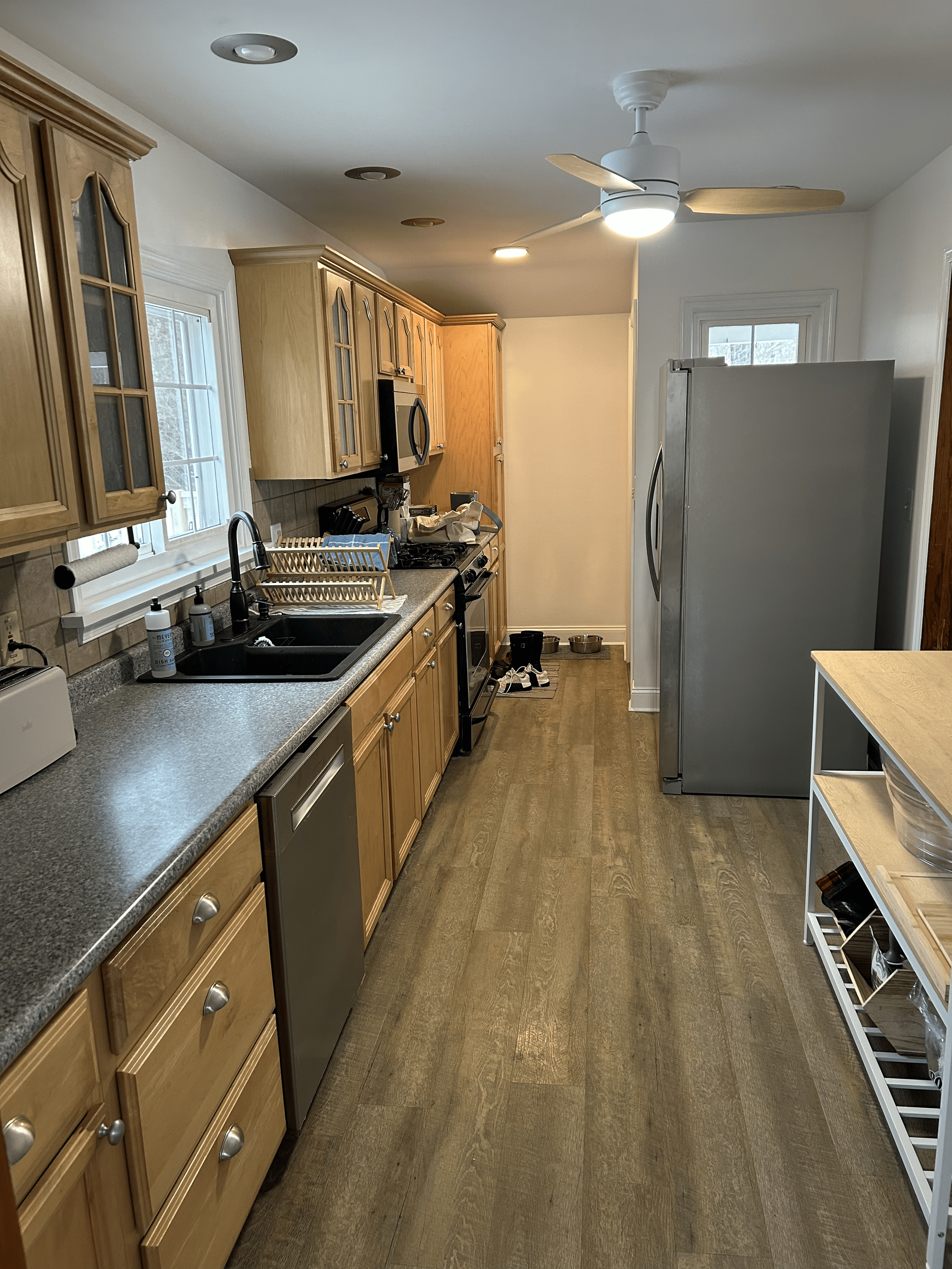 View of a kitchen with light wood cabinets, a gray countertop, a black sink, a stainless steel refrigerator, a black stove, a microwave, a white shelf, and a ceiling fan with lights. There are windows above the sink, and items on the countertop including soap bottles, a dish rack, and small appliances. Shoes are on the floor near the back, with a small mat and pet bowls.