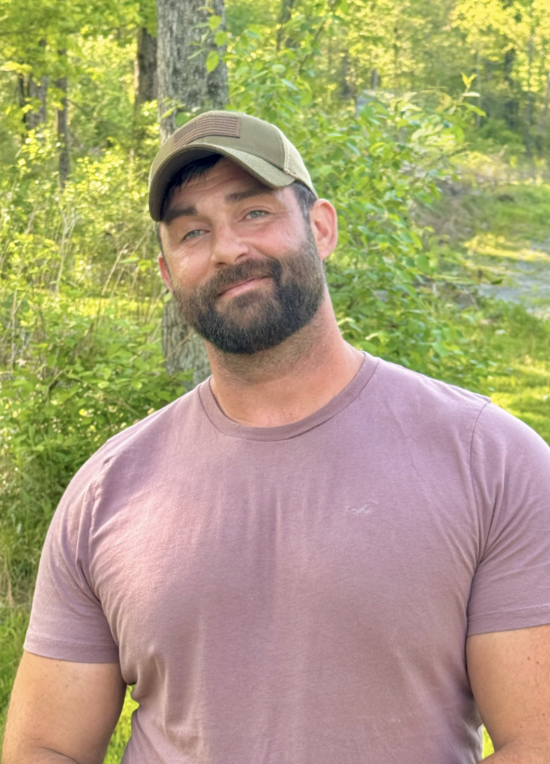 A man with a beard wearing a tan baseball cap and a mauve t-shirt, standing outdoors in a forested area with trees and greenery in the background.