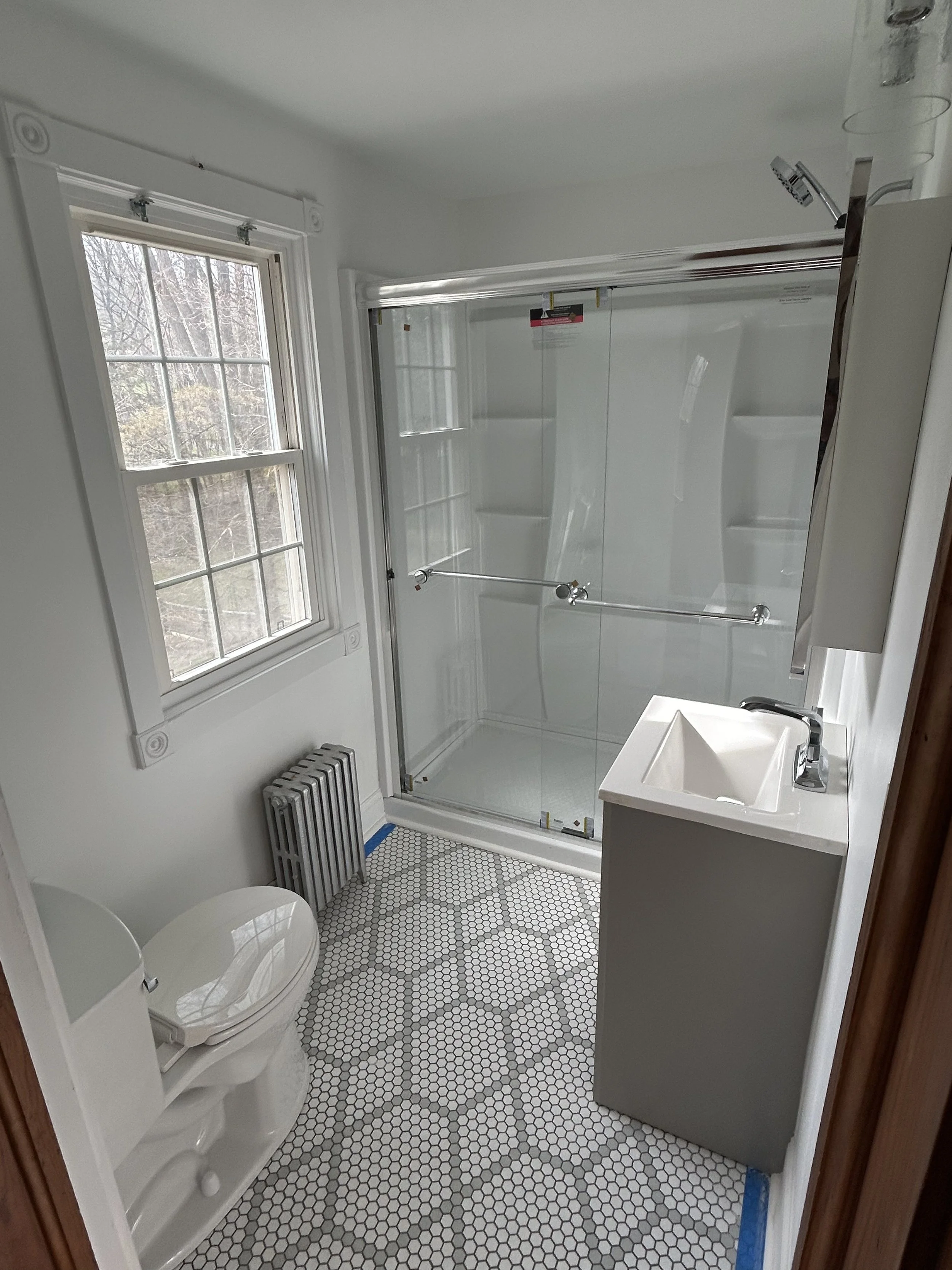 A small bathroom with white walls and hexagonal black and white tile flooring. It features a toilet, a small vanity with a white sink, a window with a white frame, and a walk-in shower with clear glass doors and a chrome showerhead.