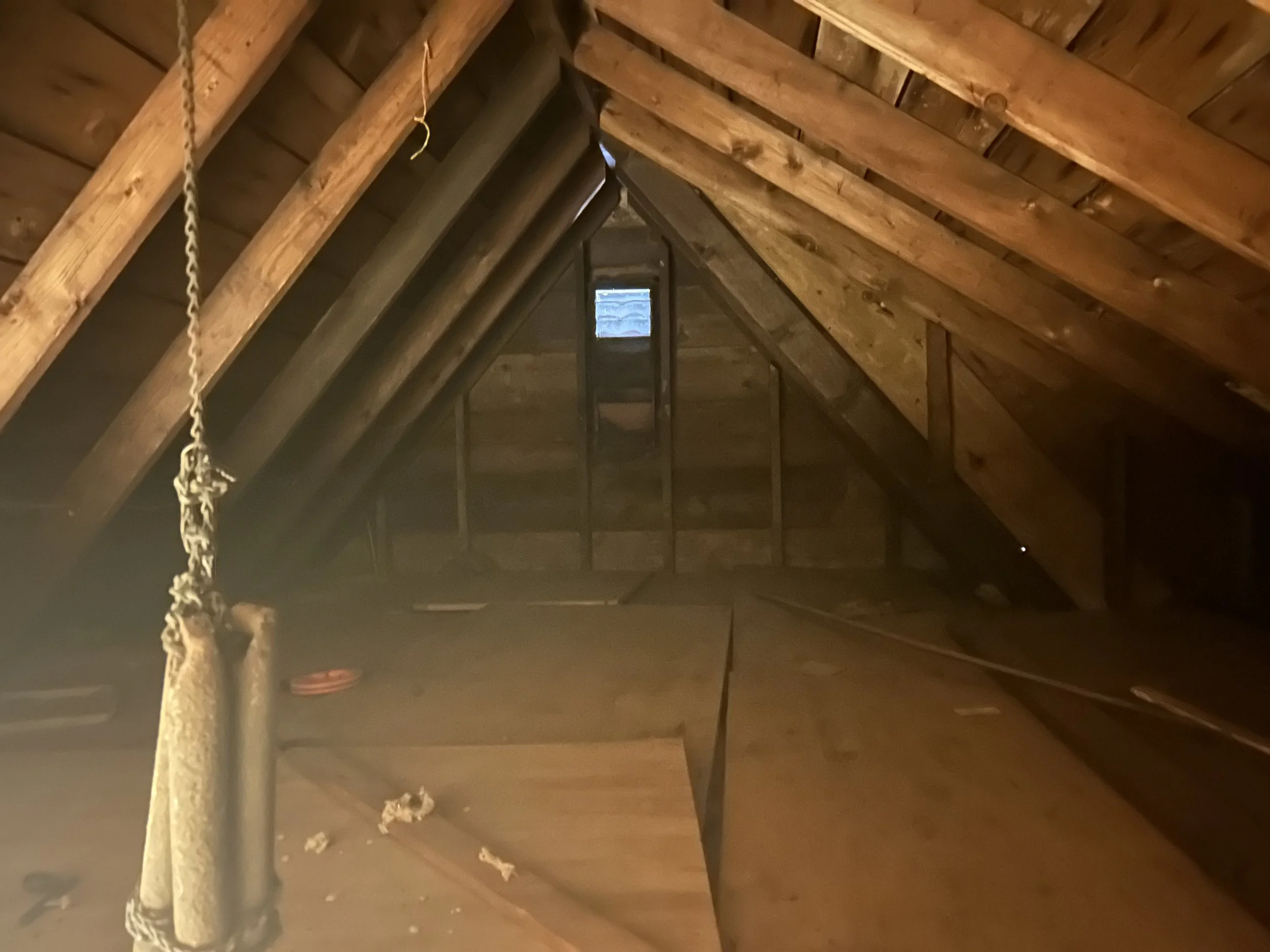 View of a house attic with unfinished wooden beams, small window at the ceiling, and some construction debris on the floor.