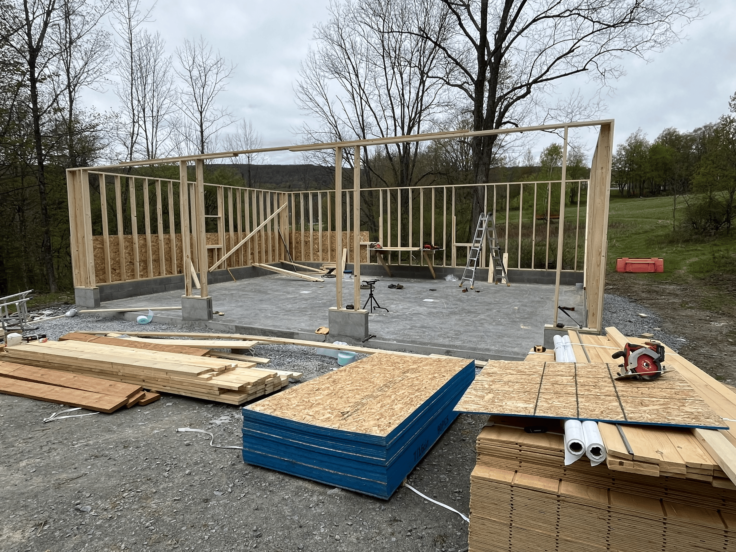 Construction site with wooden framing for a building, tools, and building materials scattered around, with a background of leafless trees and cloudy sky.