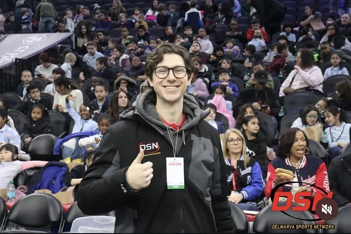 Smiling young man wearing glasses, a black jacket with a 'DSN' logo, and a lanyard, standing in front of a large crowd seated in stadium seats, with people of various ages, some holding snacks and phones, at an indoor event.