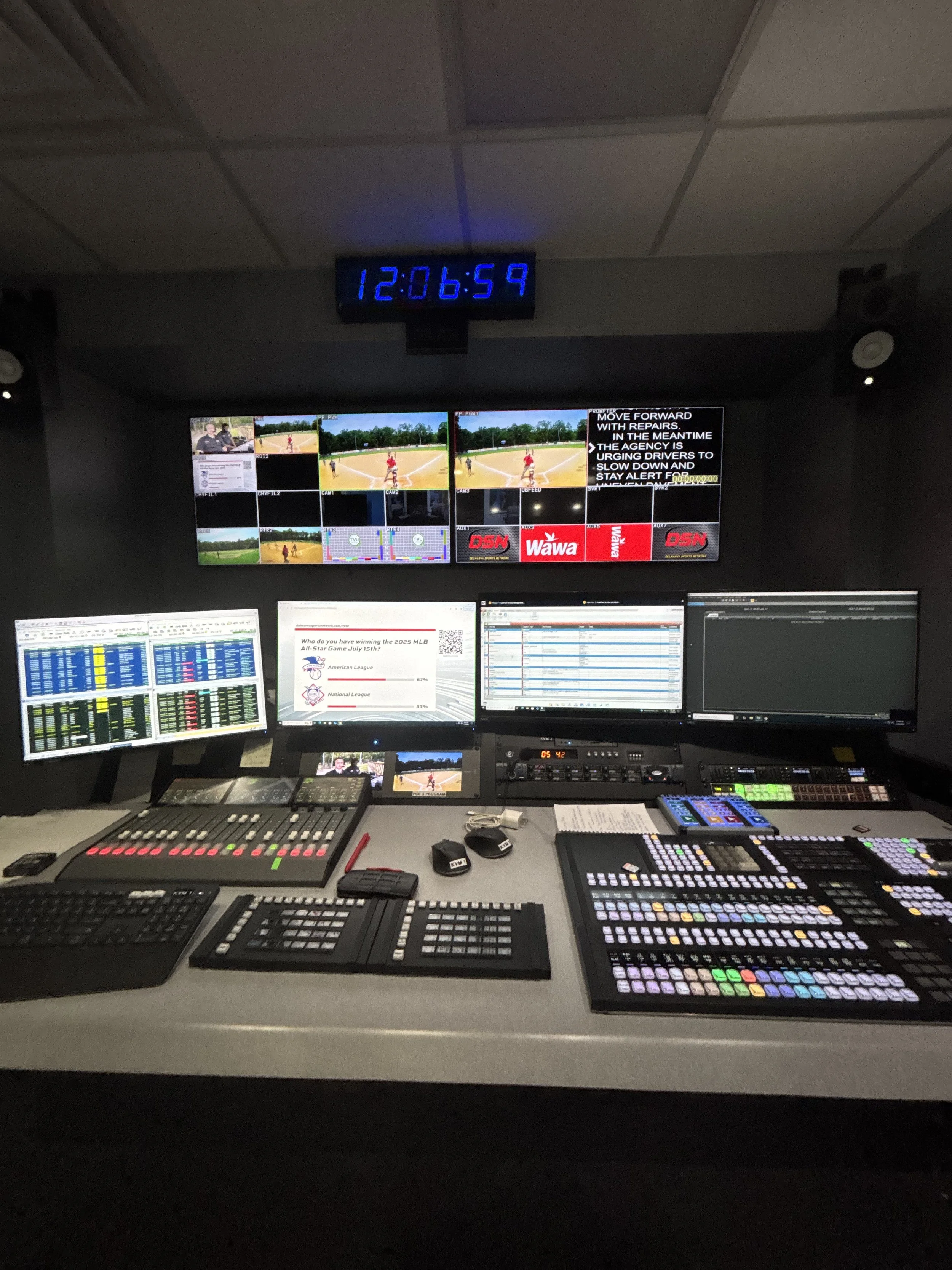 A broadcast control room with multiple computer monitors, video screens showing baseball game footage, various control panels, keyboards, and a large digital clock displaying 12:06:59.