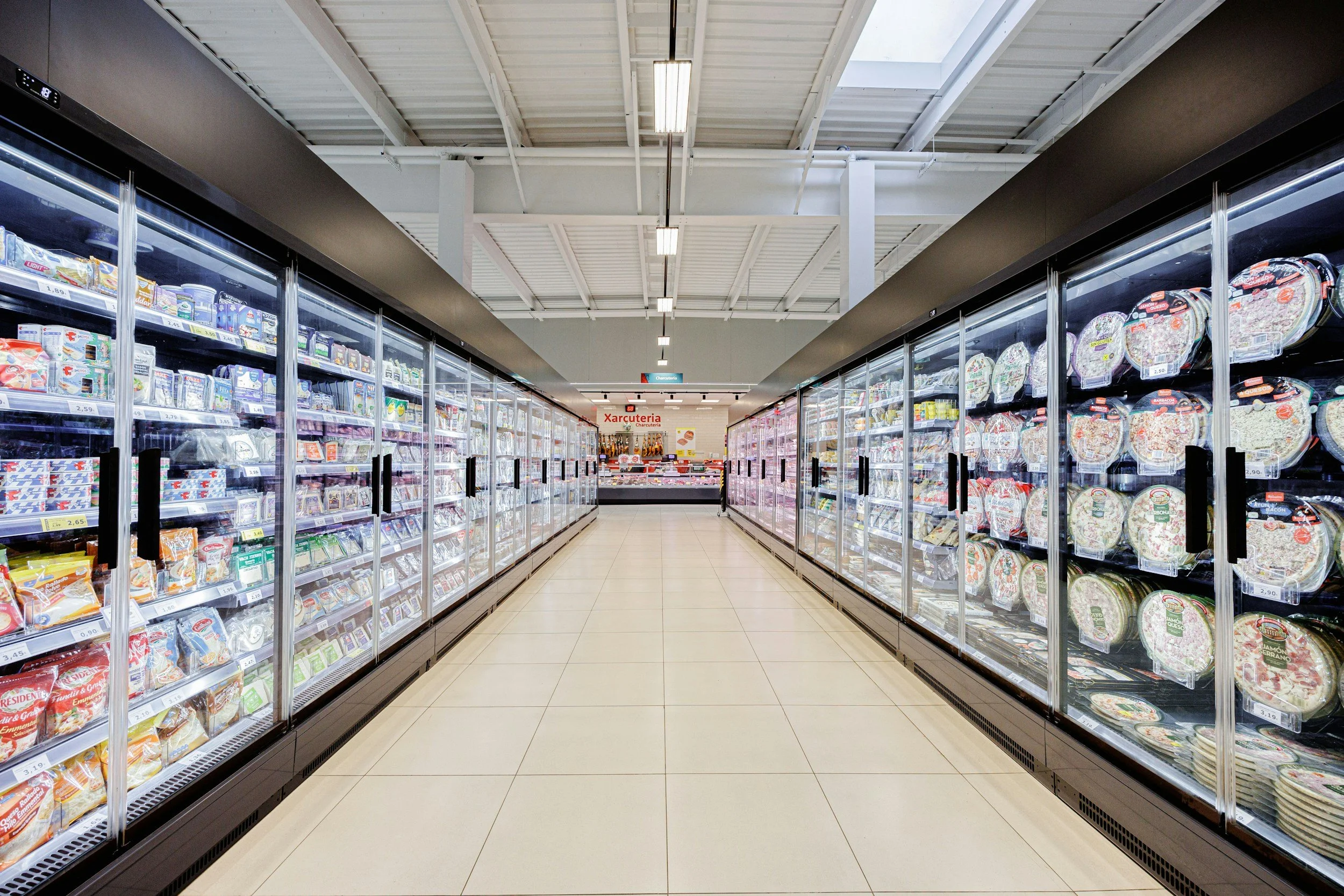 Aisle in a grocery store with frozen food section, glass freezer doors on both sides, and white tiled floor.