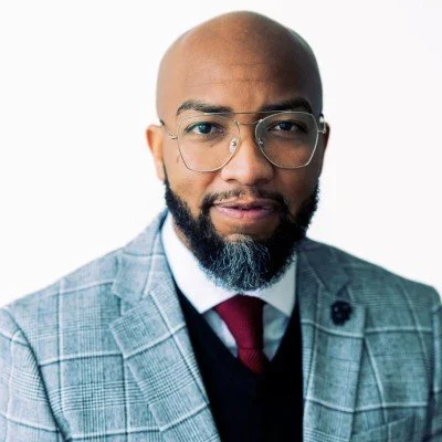 Portrait of a bald man with glasses, a beard, wearing a gray plaid suit, white shirt, and red tie, against a white background.