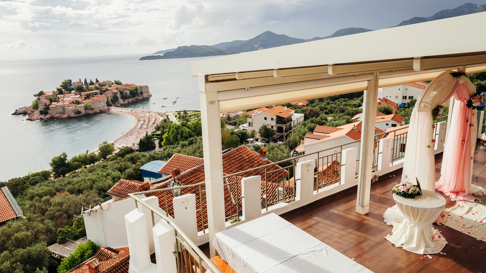 Balcony overlooking a coastal scene with a small island, beach, and mountains, decorated for a wedding with pink and white drapery and flowers.