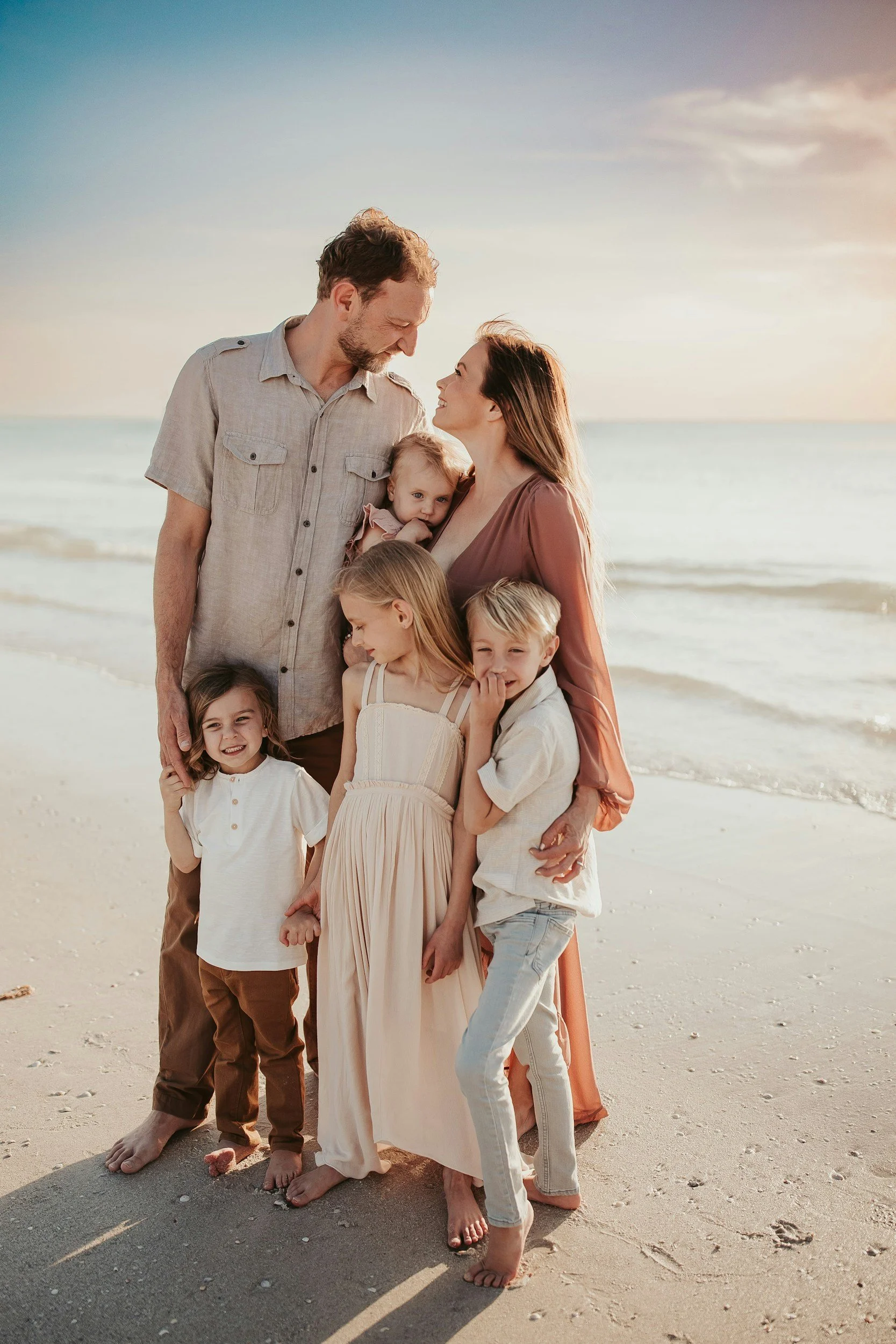 A family of seven standing on a sandy beach, with the ocean and sky in the background. The family includes two adults and five children, all smiling and looking at each other, dressed in casual light-colored clothing, barefoot.