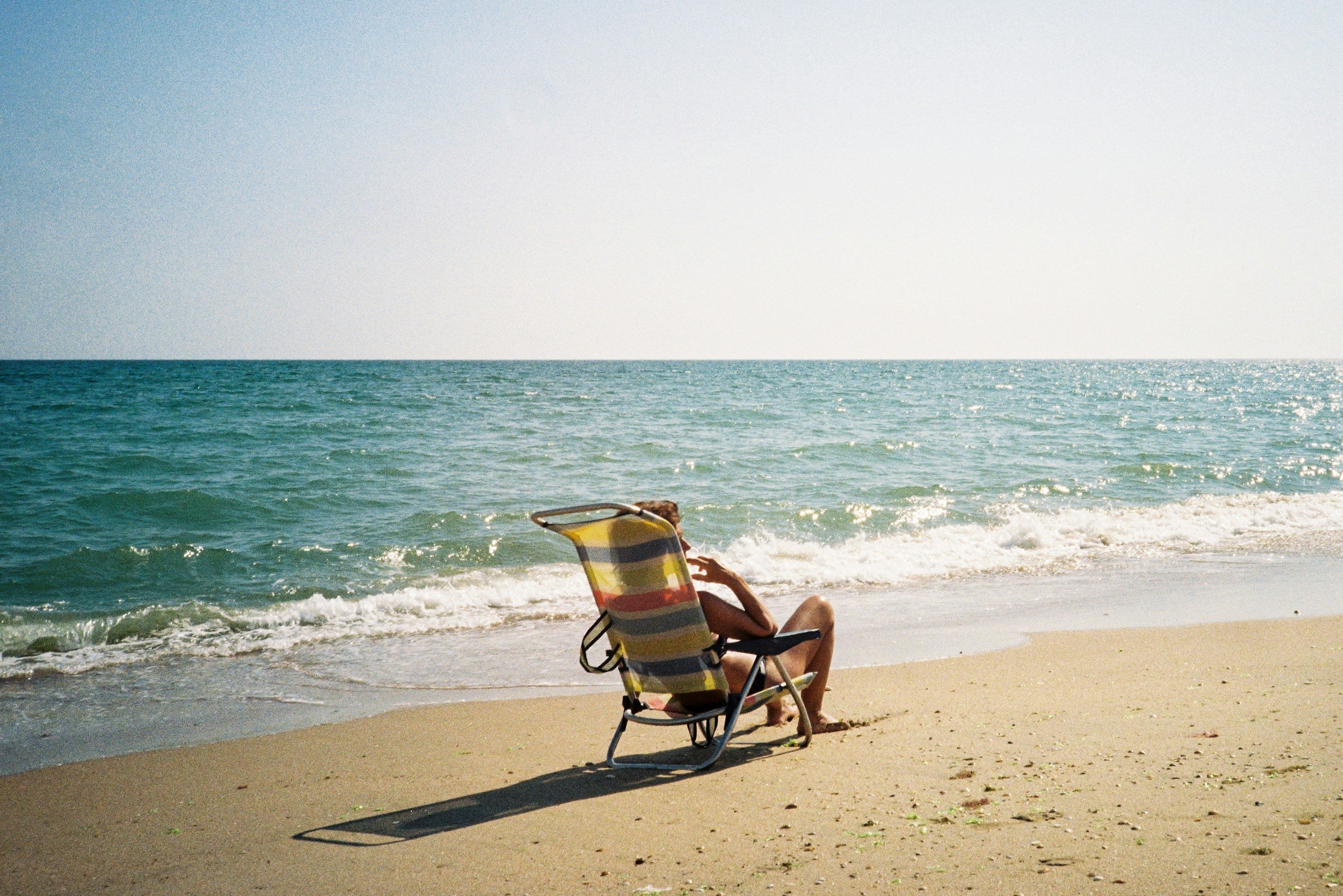 Person relaxing on a beach chair under the sun facing the ocean, with waves and a clear sky in the background.