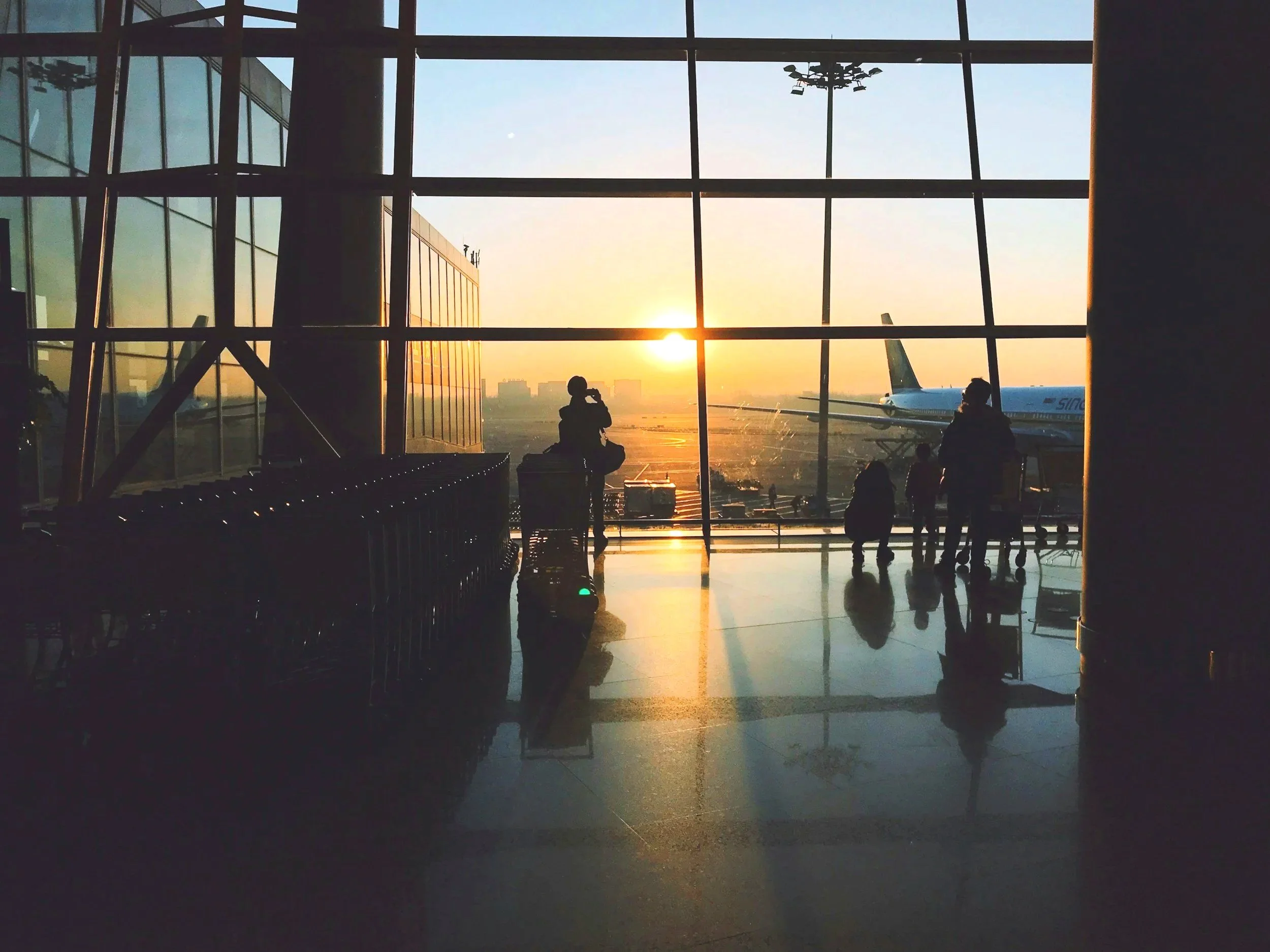 Sunset view through large glass windows of an airport terminal showing an airplane on tarmac and people sitting or standing inside.