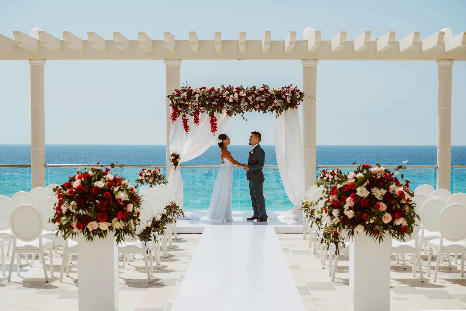 A wedding ceremony taking place outdoors on a balcony with a view of the ocean. A bride and groom are holding hands under a floral arch. The setting includes white chairs and decorated tables with red, pink, and white flowers.