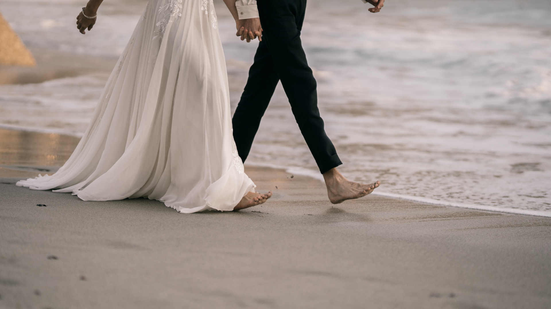 A couple dressed in wedding attire walking barefoot on the beach by the shoreline, holding hands.