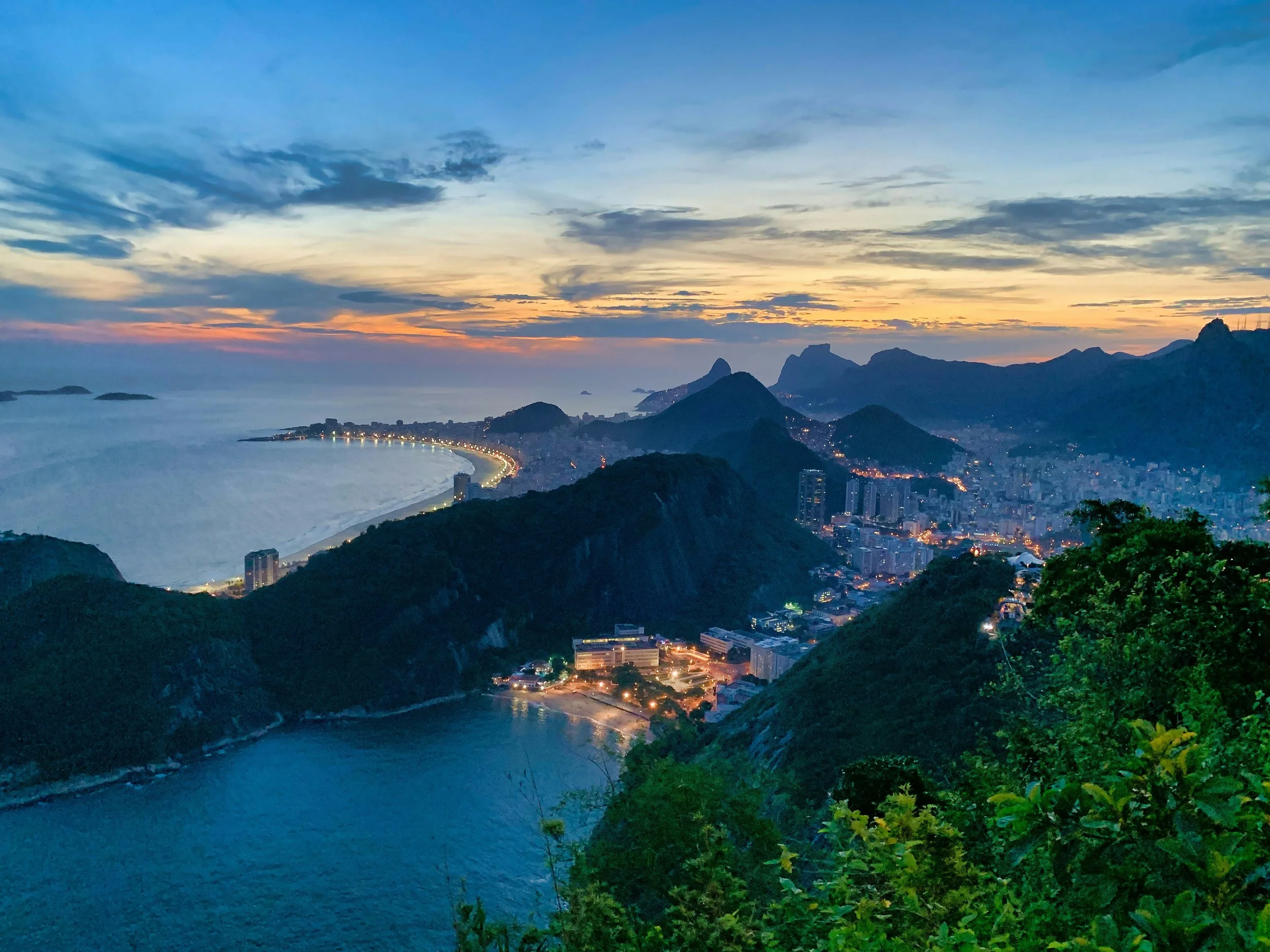 A scenic view of a coastal city during sunset, with mountains in the background and a curving beach in the foreground. The sky displays shades of blue, orange, and yellow, and city lights are beginning to turn on.