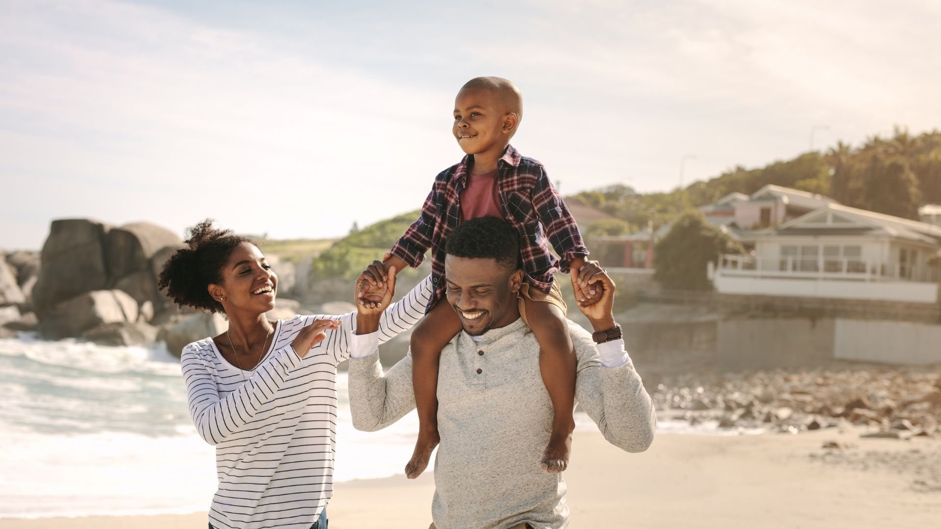 A family enjoying a day at the beach with a woman, a man, and a boy riding on the man's shoulders, smiling and playing together near the water with houses and rocks in the background.
