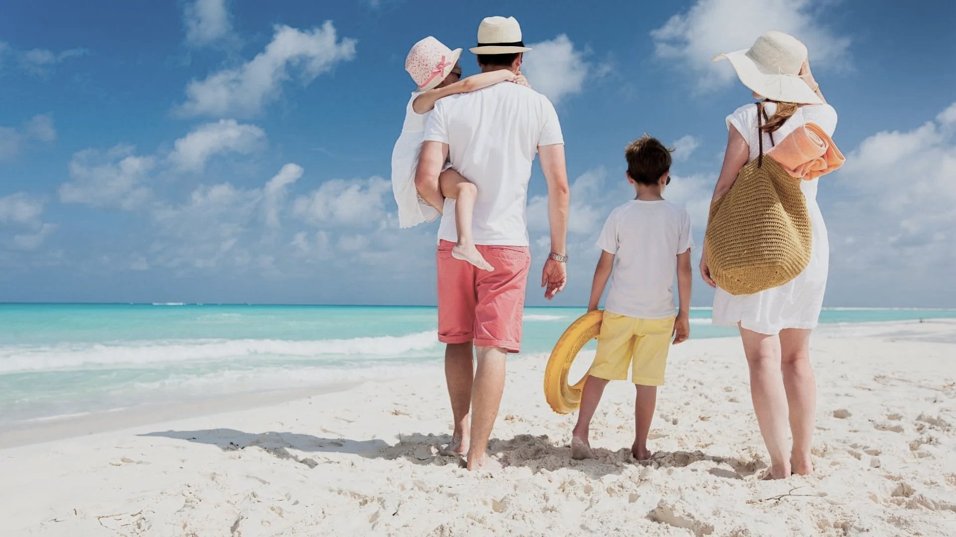 A family walking on a sandy beach near the ocean under a partly cloudy sky. The father carries a young girl on his back, and the mother and two boys walk beside him, carrying a beach float and a rolled-up towel.