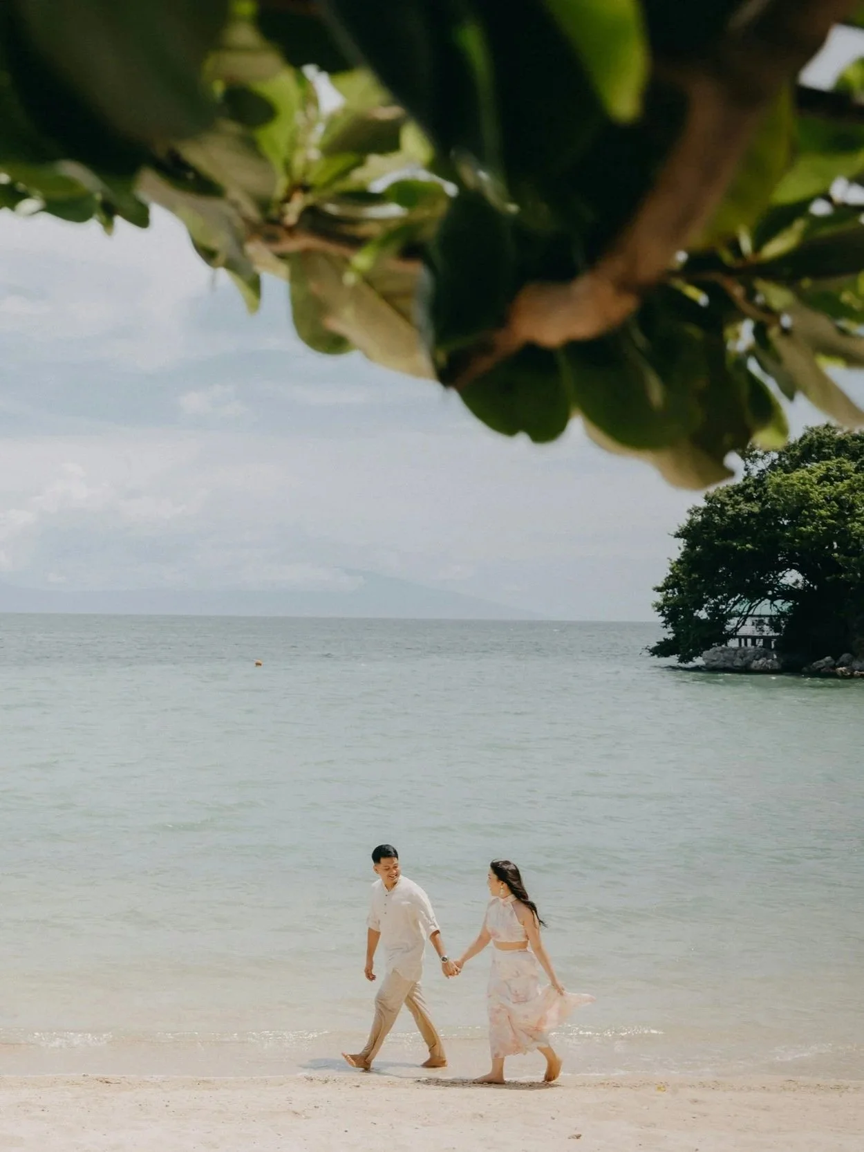 A couple walking hand in hand along the beach near the ocean, with trees in the background and a cloudy sky above.
