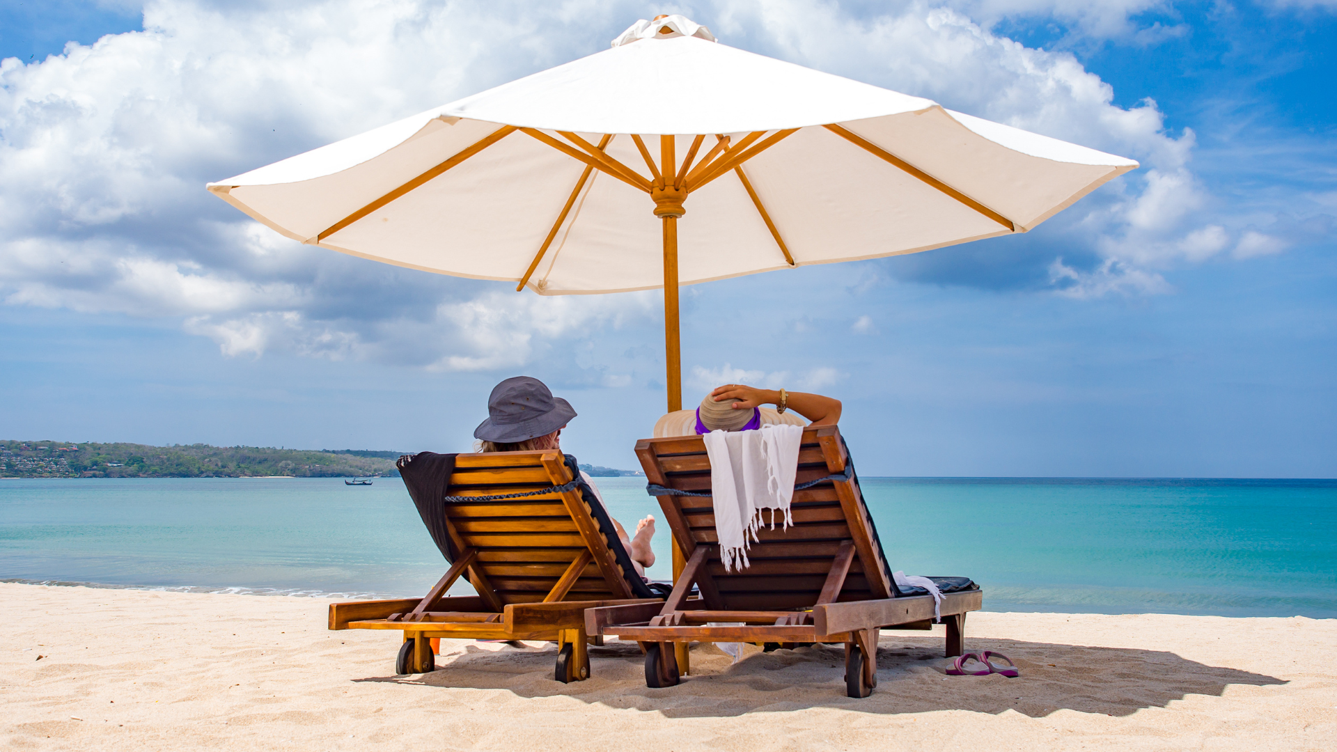 Two women sitting on wooden lounge chairs under a large white beach umbrella on a sandy beach, facing the ocean with a distant shoreline and blue sky with clouds.