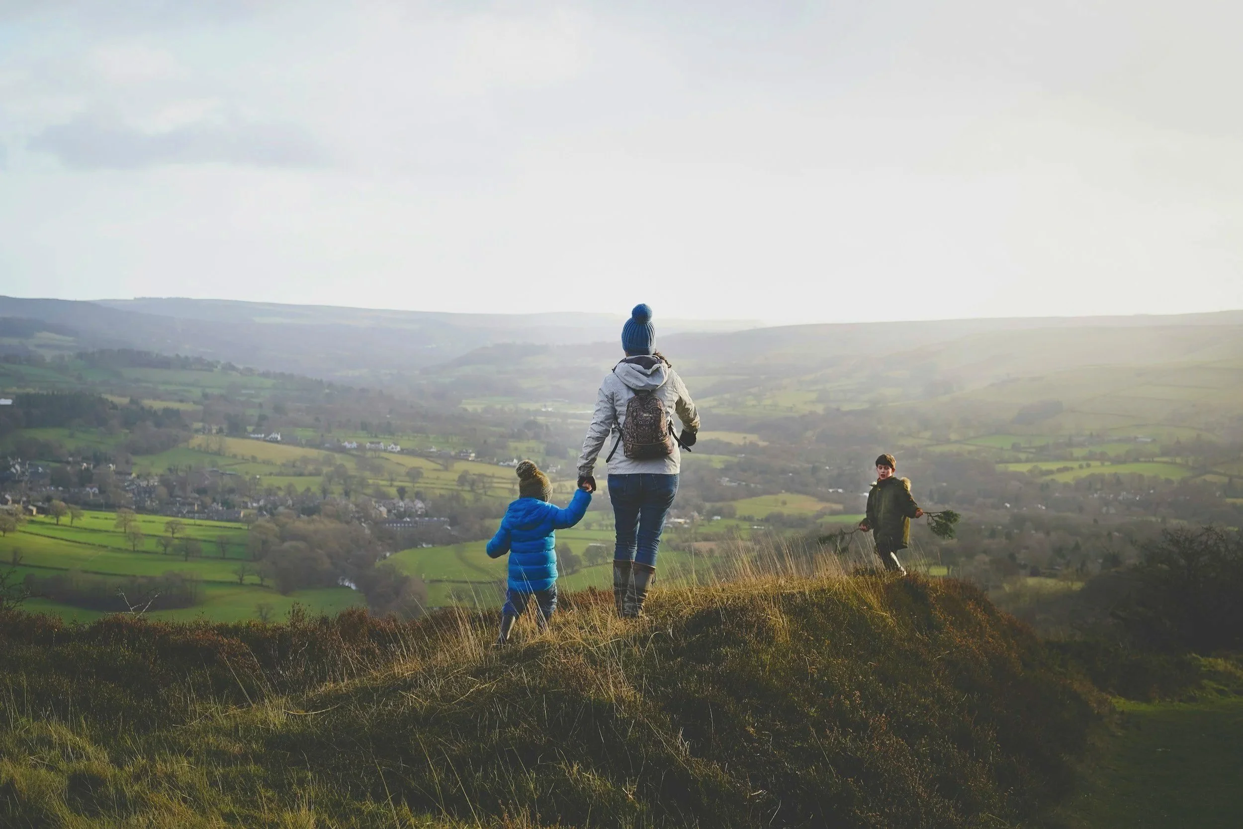 A woman holding hands with two children while walking on a grassy hilltop overlooking a valley with houses and fields, under a partly cloudy sky.