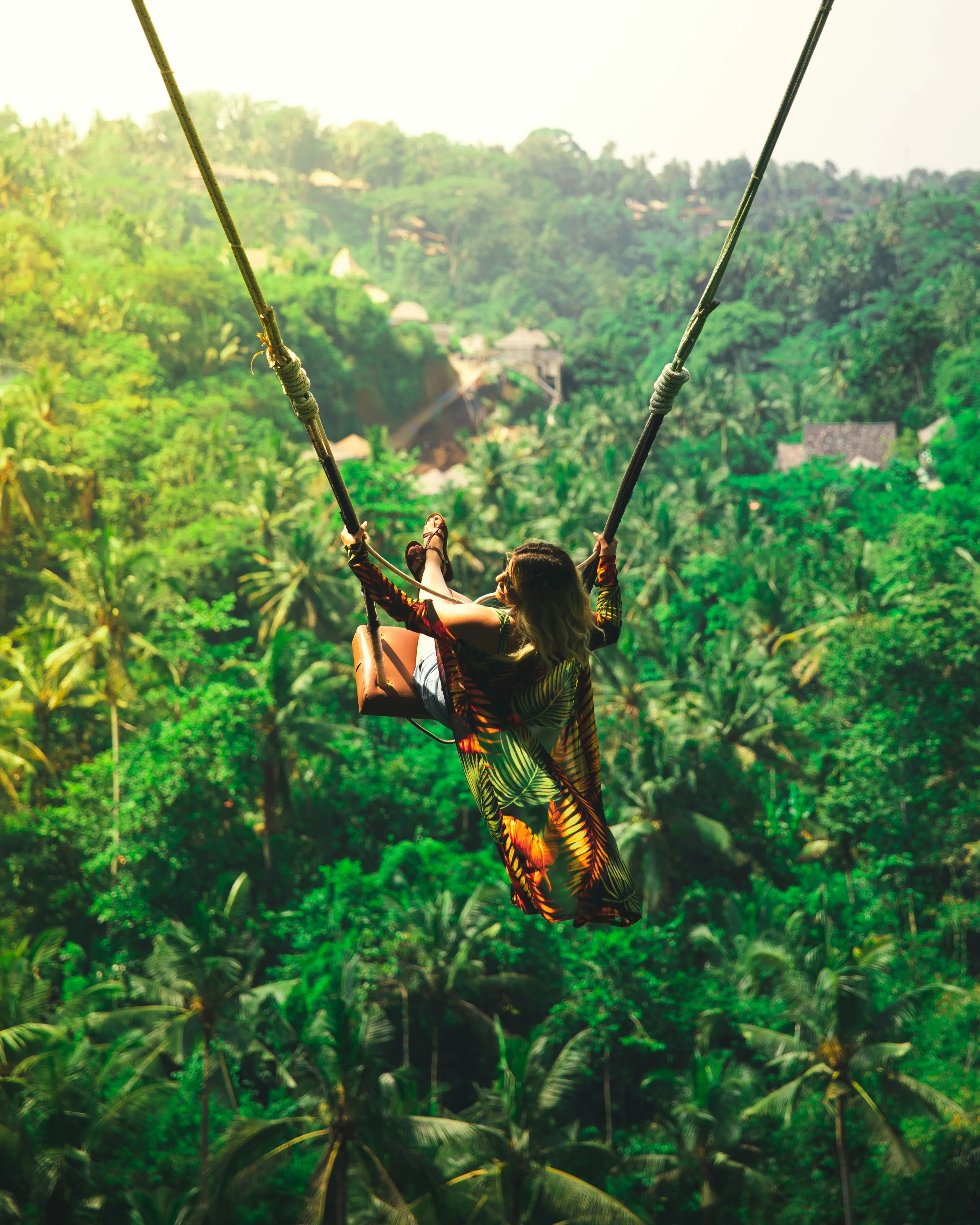 Person on a swing in a lush green forest, with hilltop houses visible in the background.