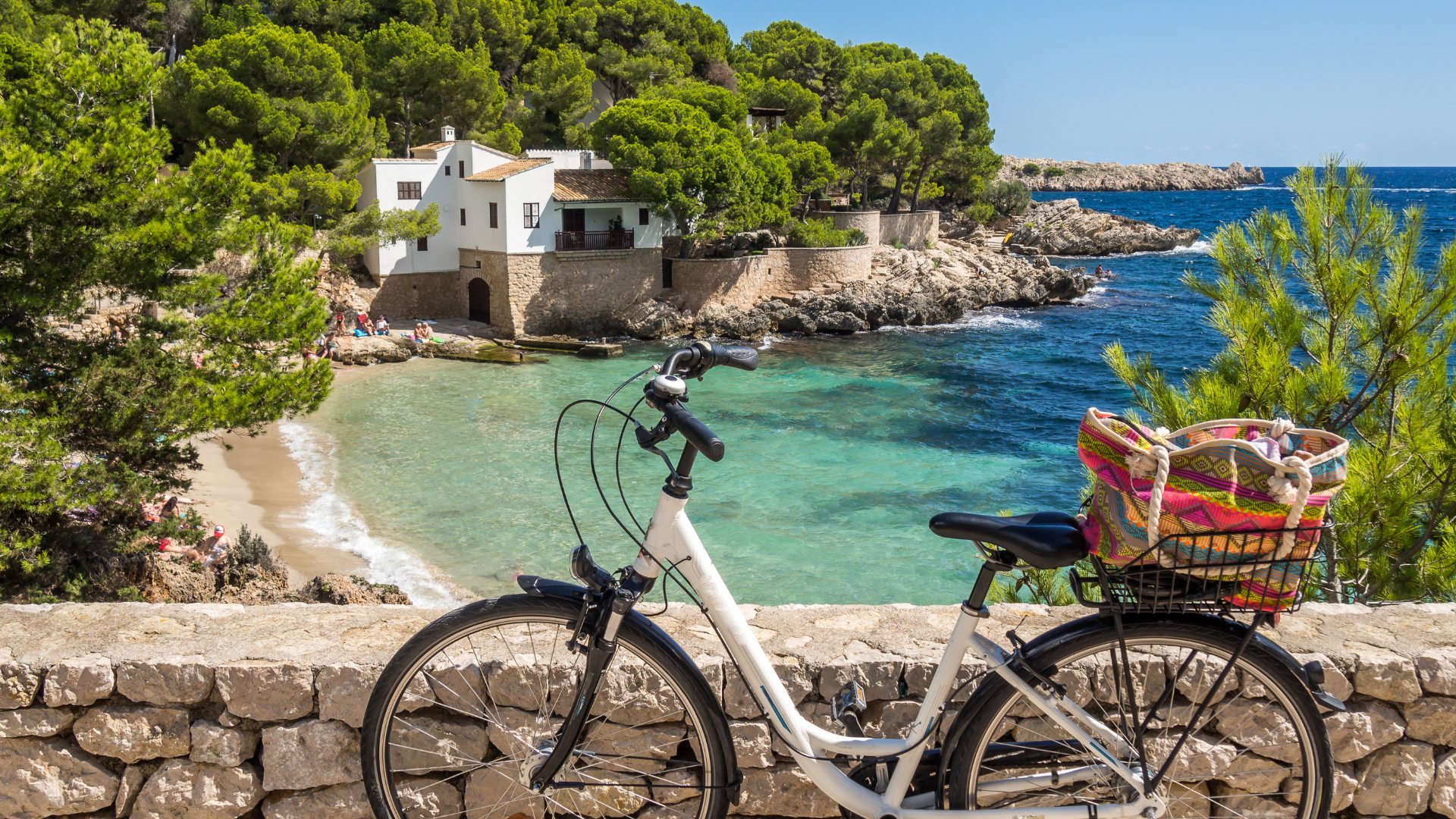 A white bicycle with a black seat and a colorful woven basket attached to the rear, parked in front of a stone wall along a coastal scene. Behind it, there is a beach with people sunbathing, turquoise waters, rocky cliffs, lush green trees, a white house on the hillside, and the blue sea under a clear sky.
