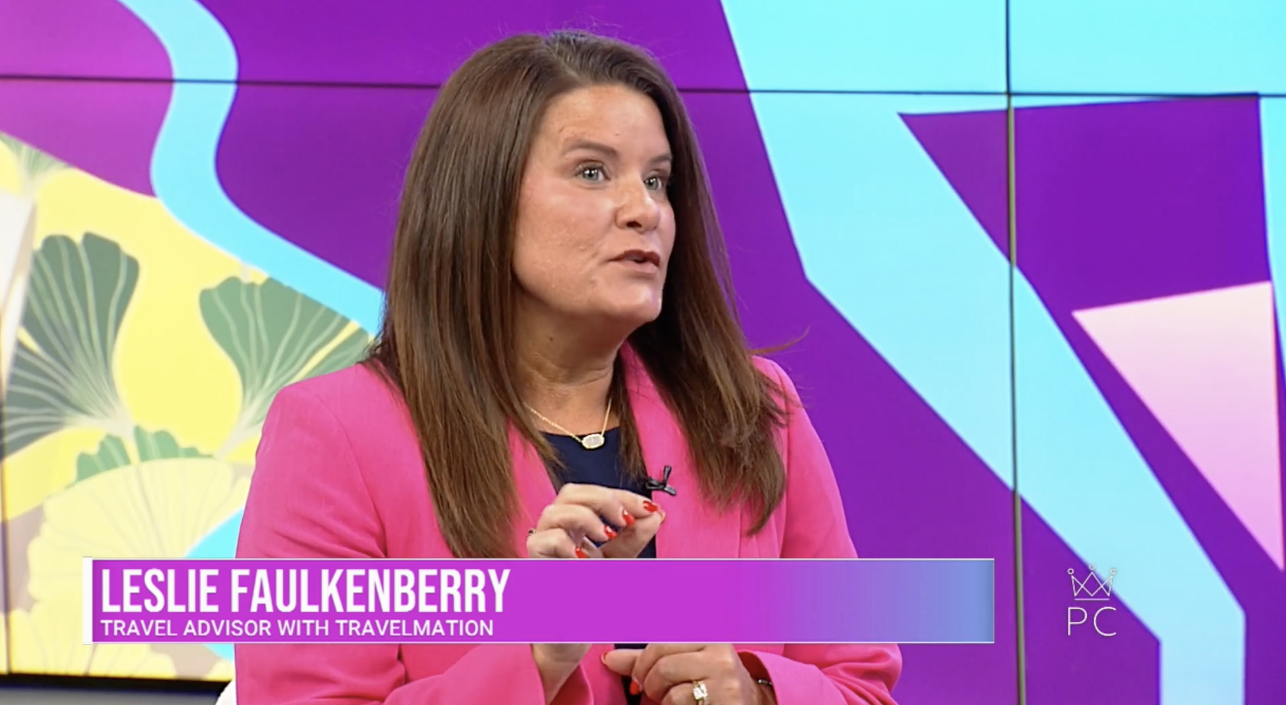 Leslie Faulkenberry, a woman with long brown hair, wearing a pink blazer and a necklace, speaking on a TV show. The background is colorful with blue, purple, and yellow elements.