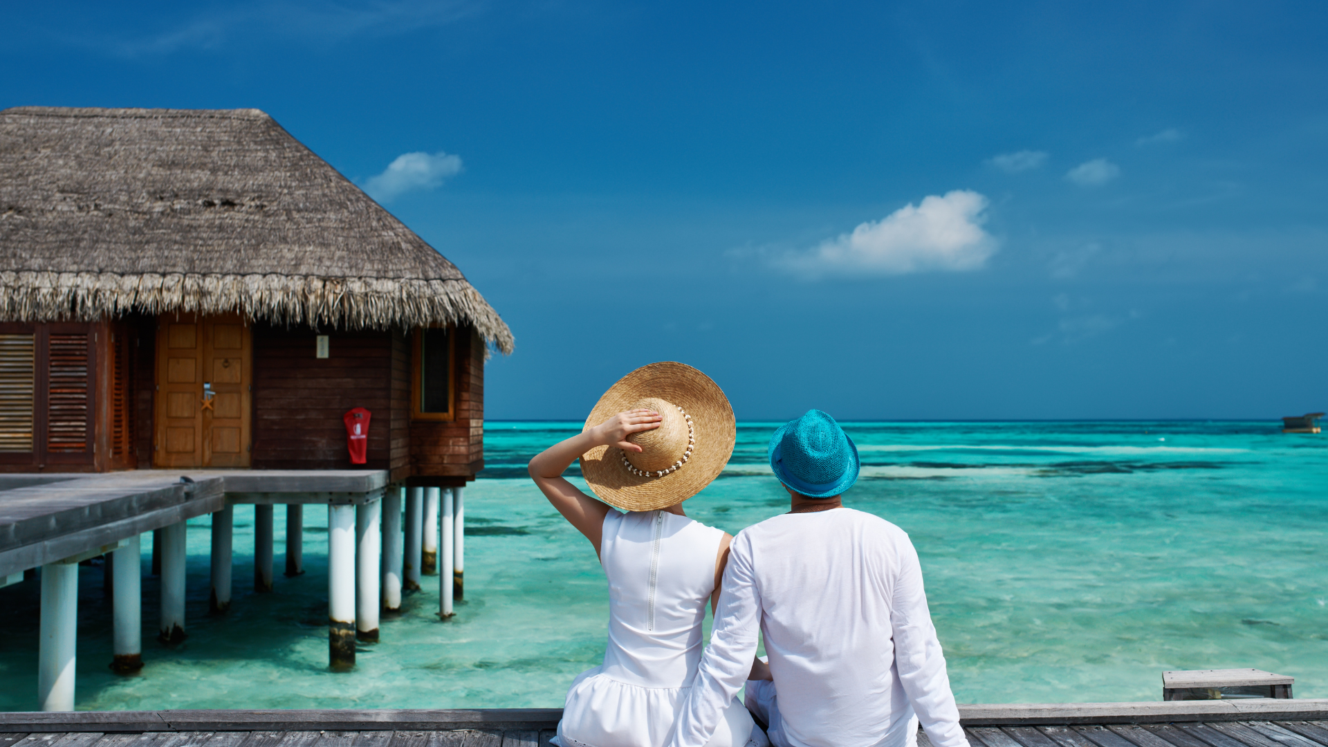 A woman and a man sitting on a wooden pier, facing over ocean water towards overwater bungalow with thatched roof, one woman covering her face with straw hat, the other pointing at the horizon, both dressed in white, with clear turquoise sea and sky with clouds in the background.