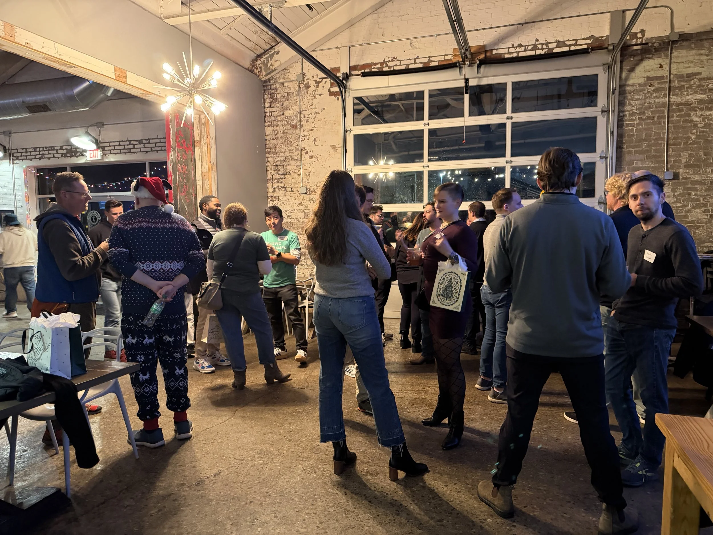 Indoor holiday gathering with people chatting, wearing casual clothes, some with Christmas-themed pajamas, in a rustic venue with exposed brick walls and large windows, decorated for Christmas with lights.
