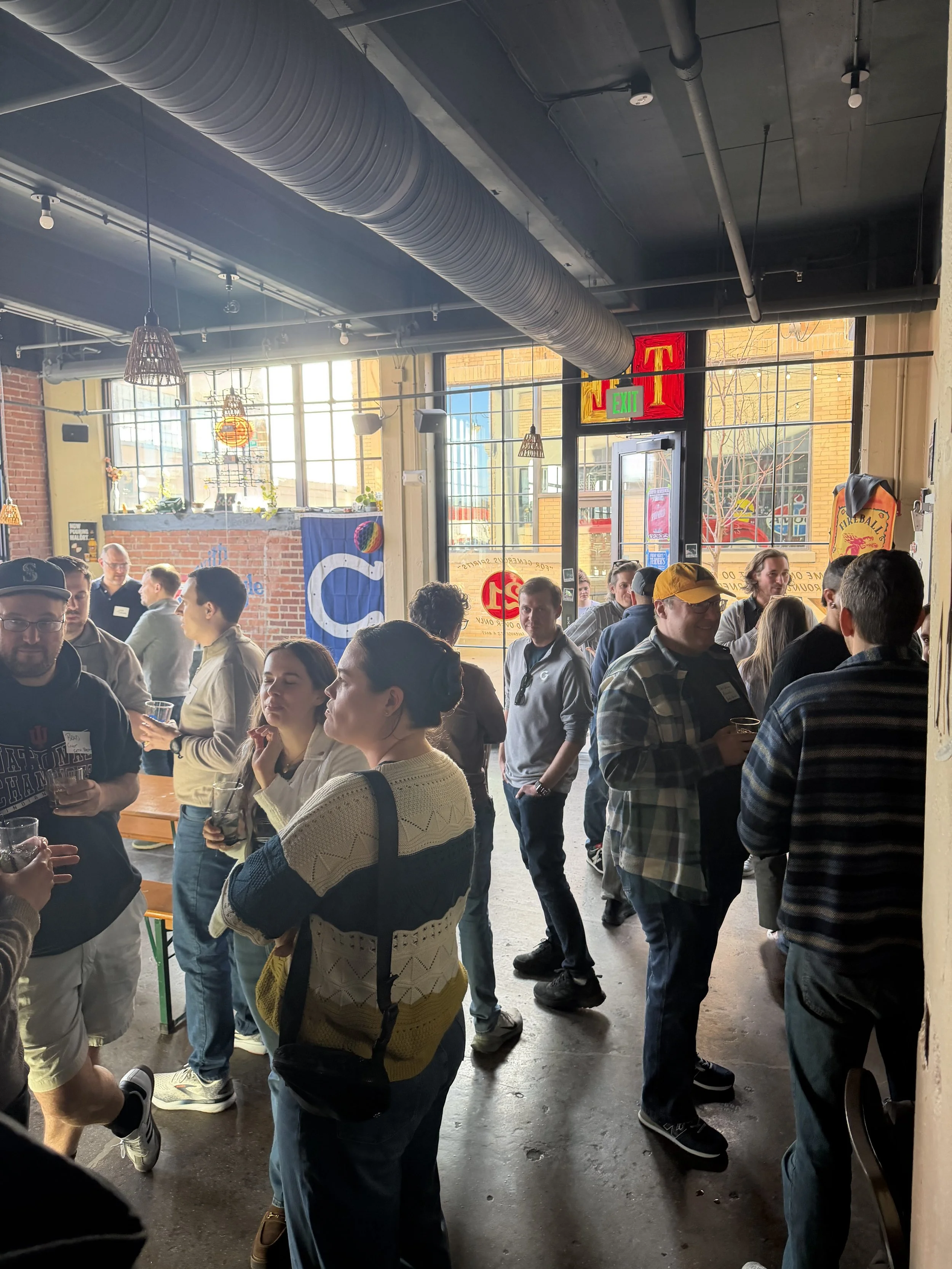 Indoor gathering with people socializing and enjoying drinks in a bright, industrial-style space with large windows and exposed ductwork.