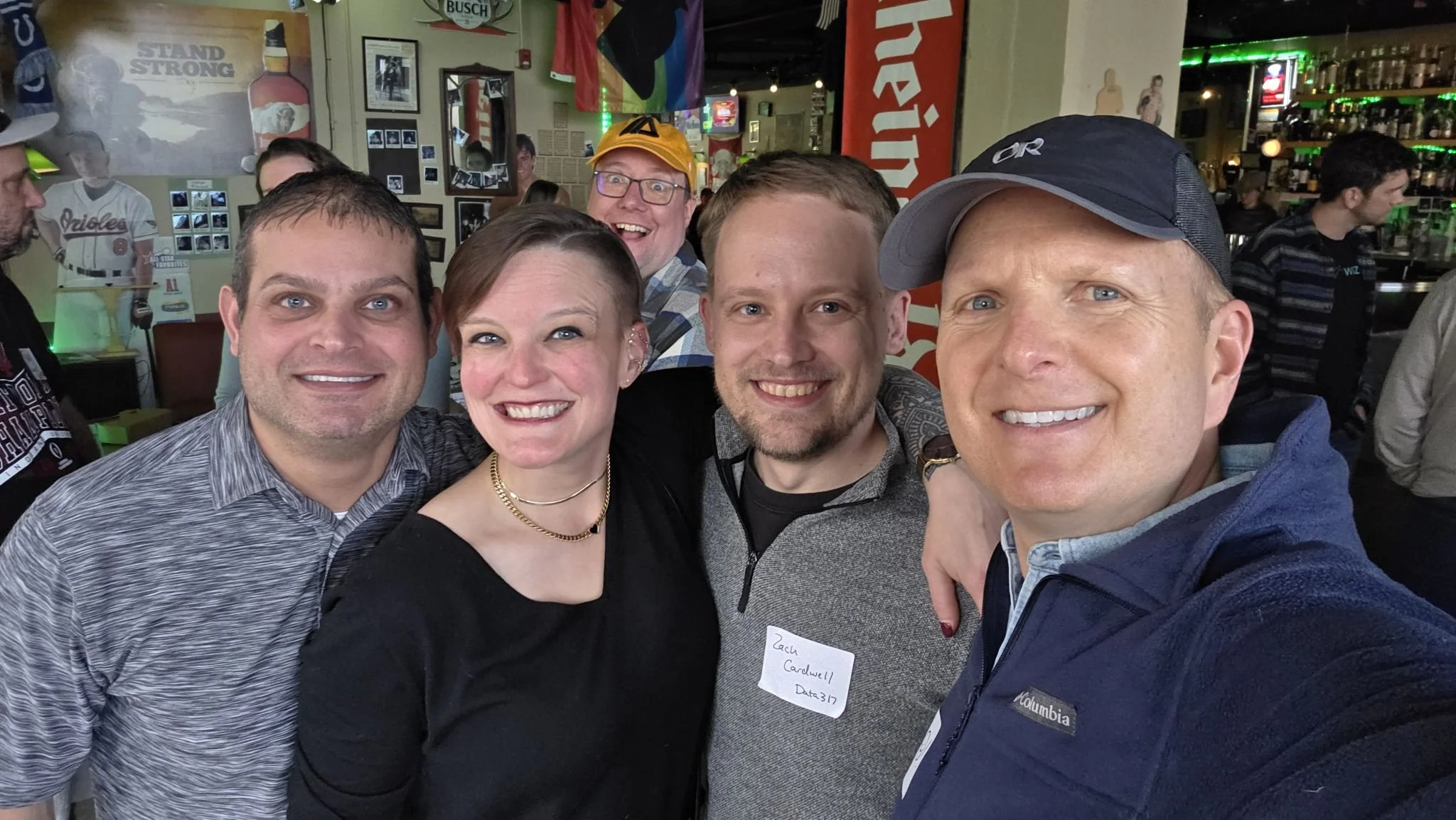 A group of six smiling people taking a selfie in a bar or restaurant. The background features framed pictures, a rainbow flag, and bar shelves with bottles and glasses.