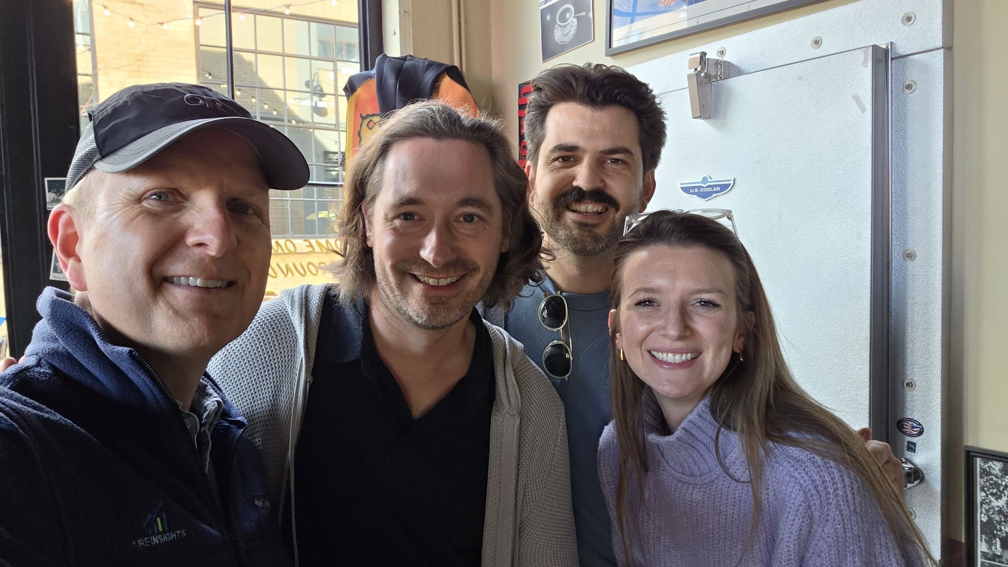 A group of five smiling people, three men and two women, taking a selfie inside a building near a window. Behind them are framed pictures and a wall with various signs.