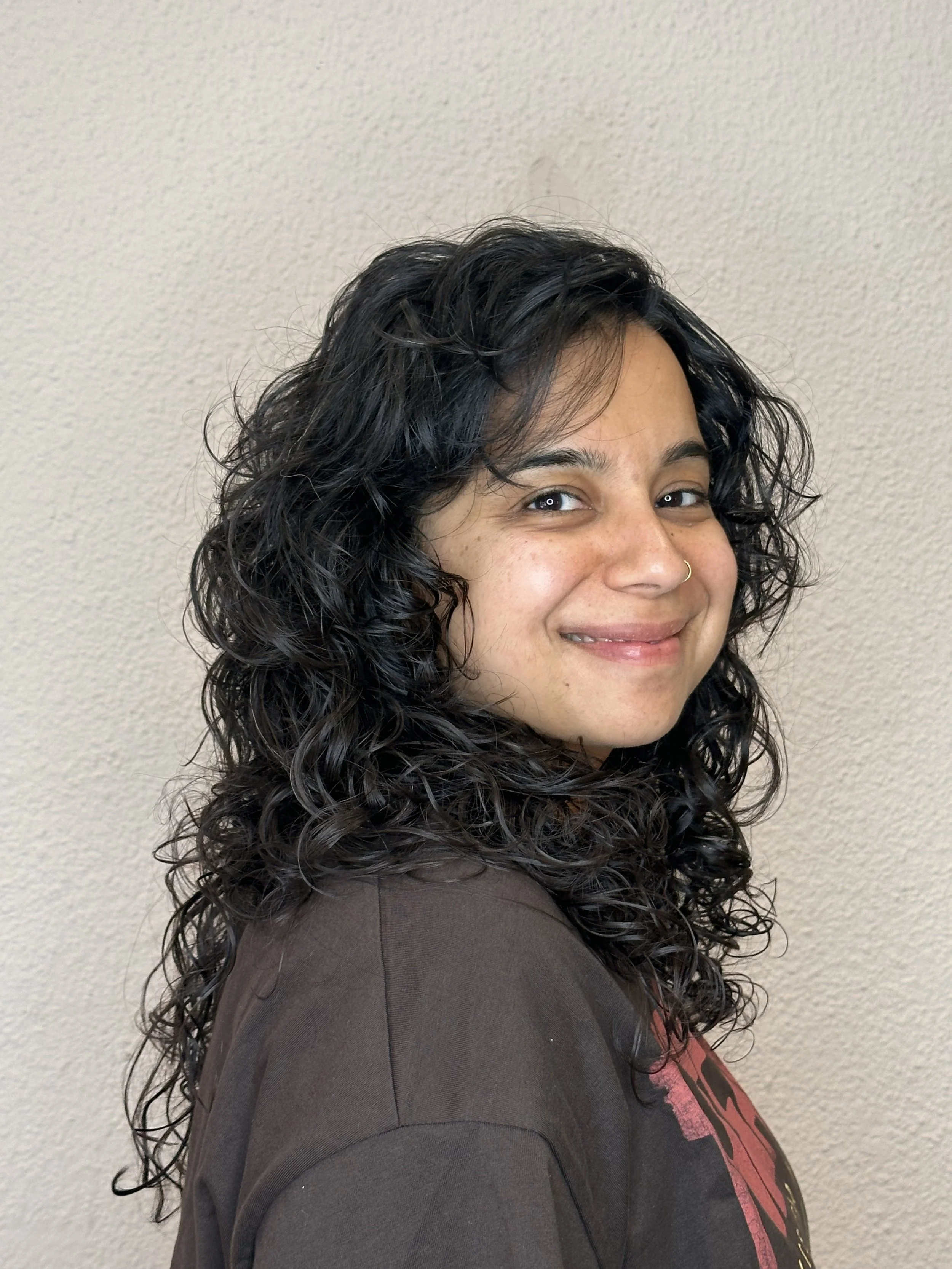 A woman with dark, curly hair smiling in front of a plain beige wall.