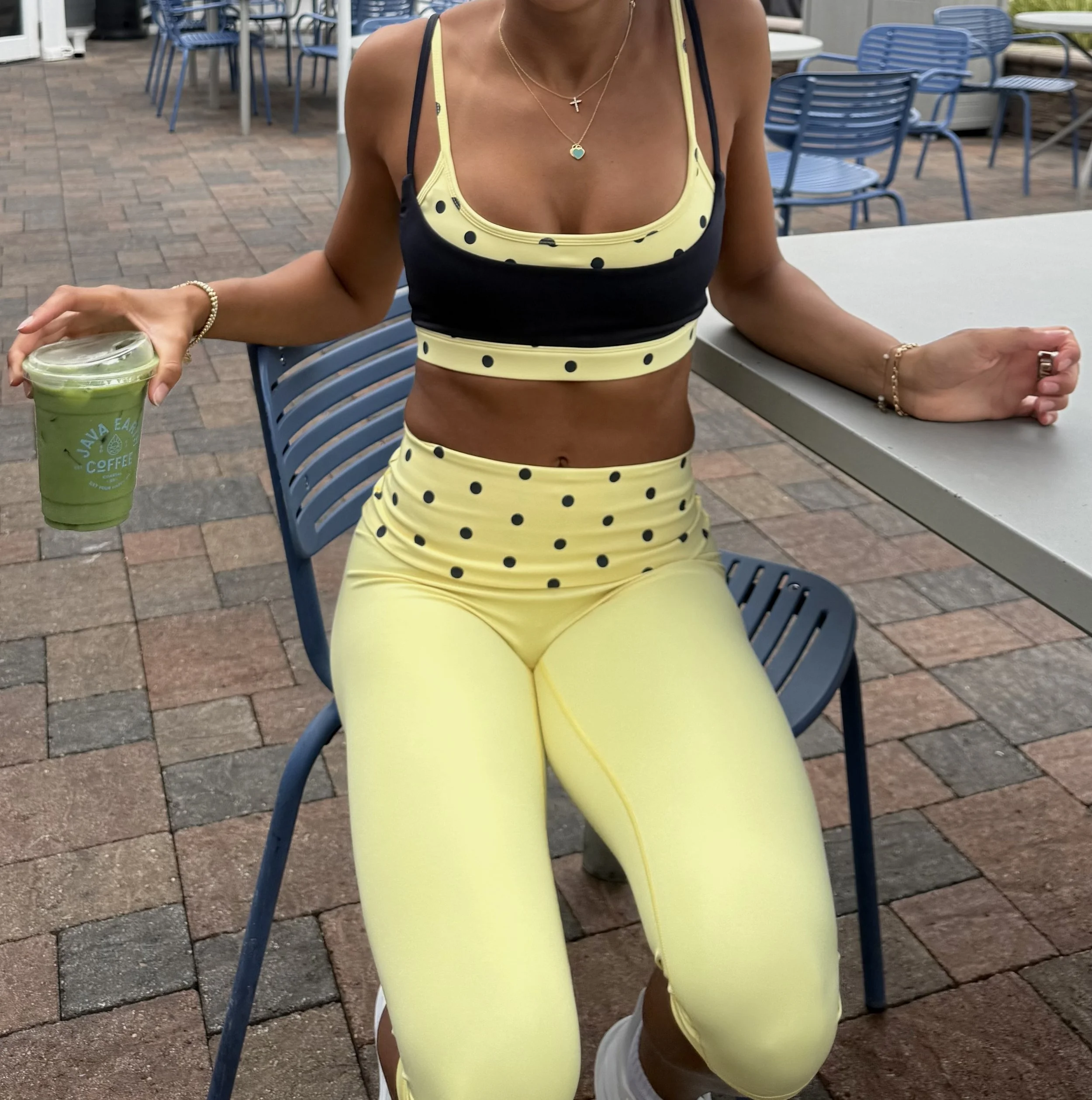 A woman wearing a yellow and black polka dot sports bra and matching leggings, sitting at an outdoor seating area with blue chairs and tables. She has jewelry, including necklaces and bracelets, and is holding a green beverage. The setting appears to be a patio with brick flooring.