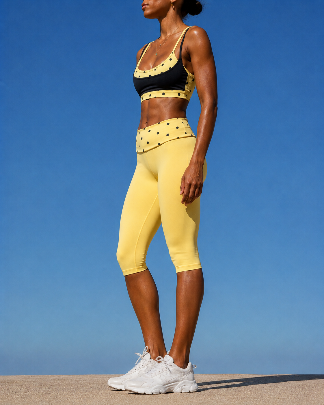 A woman wearing yellow and black activewear, standing outdoors under a clear blue sky, with her body facing slightly to the right and head turned away.
