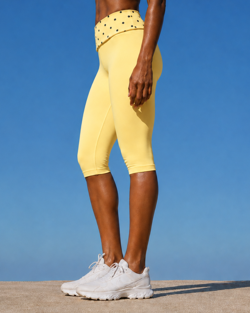 Close-up of a person wearing yellow athletic leggings with a black polka dot waistband, white sneakers, and standing outdoors against a blue sky background.