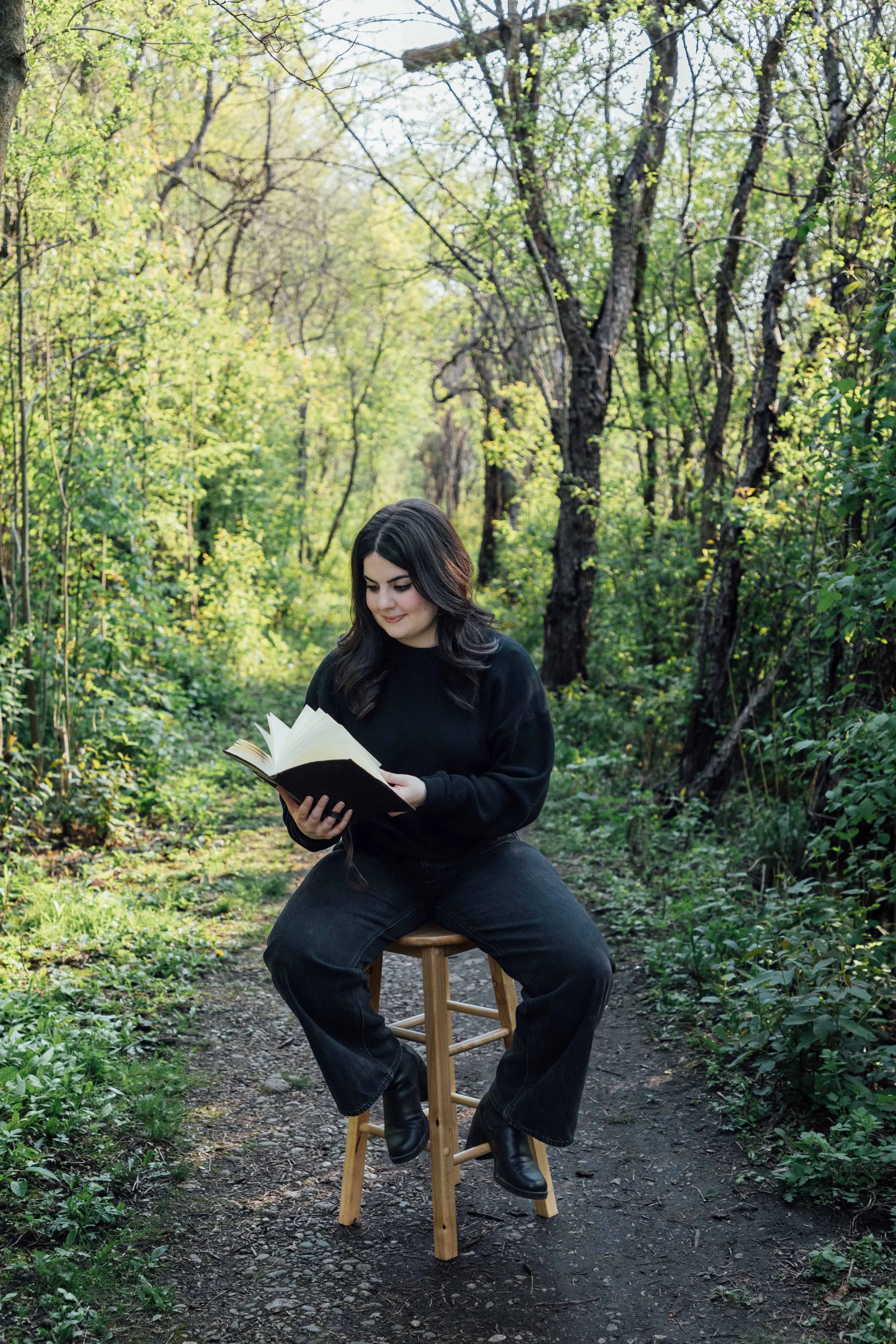 Young woman with dark hair sitting on a stool on a dirt path in a lush green forest, reading a book.