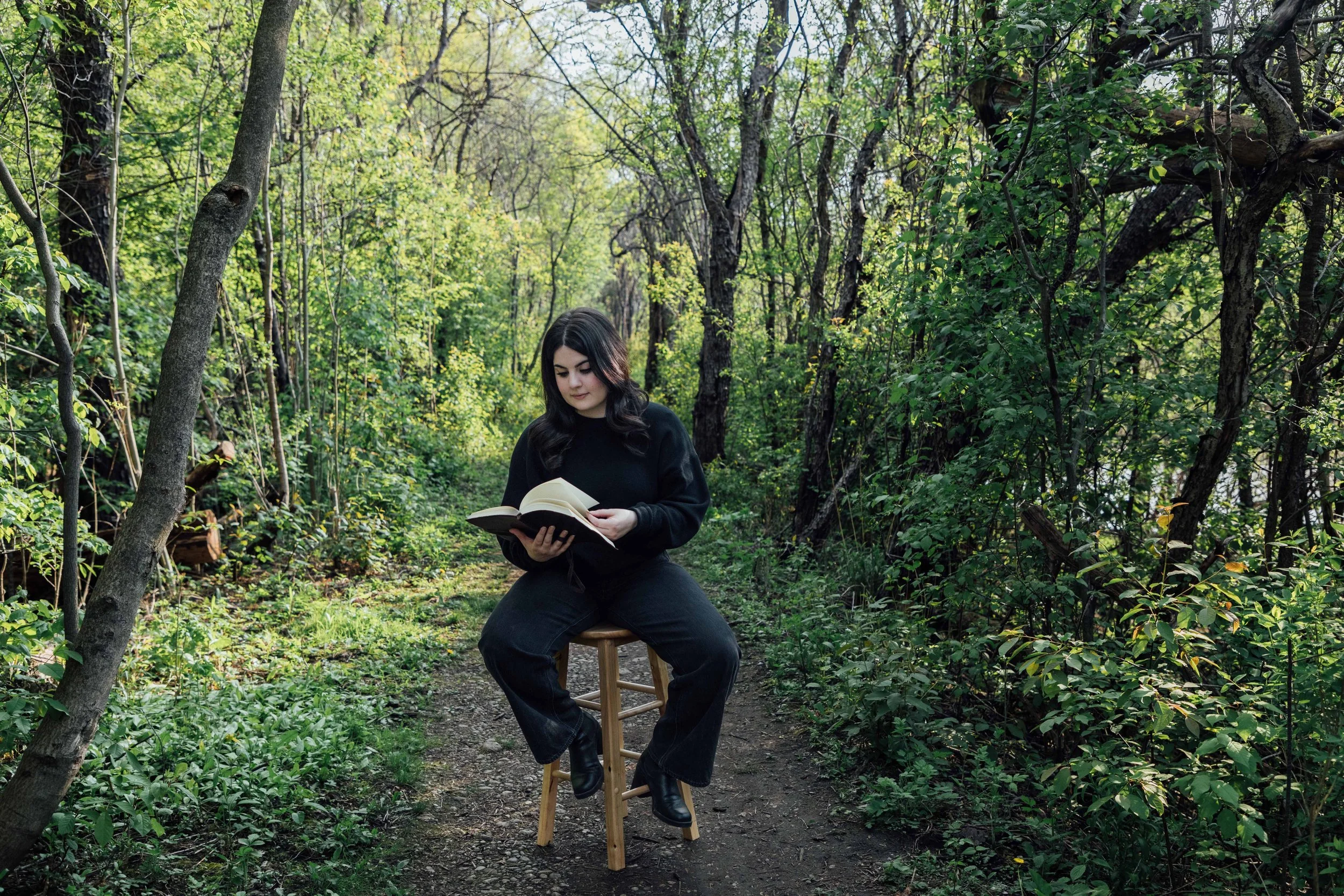 A woman with black hair dressed in black, sitting on a stool in a forest, reading a book.
