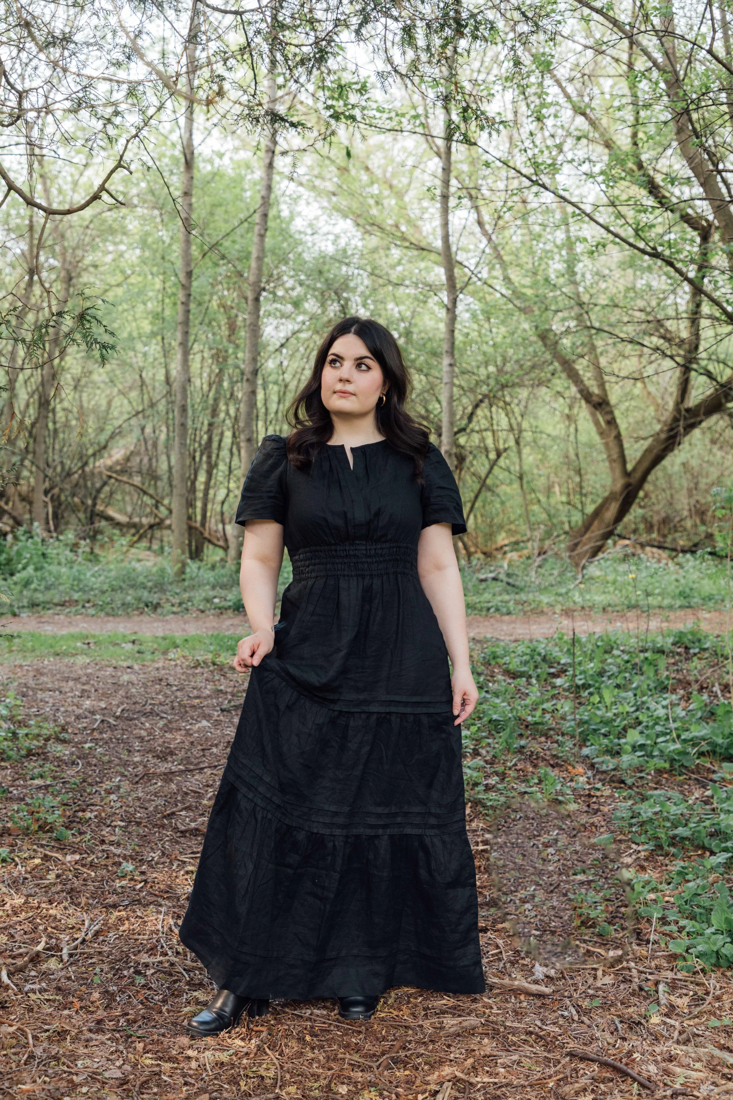 A woman in a long black dress and black boots standing outdoors in a wooded area with trees and green foliage.