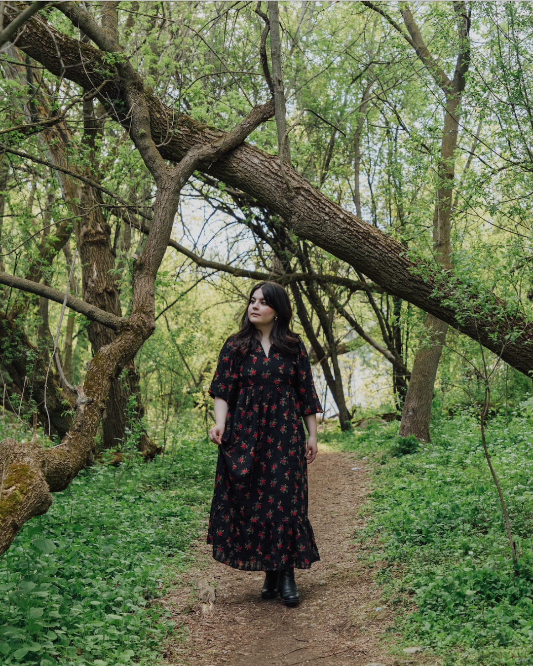 A woman in a black floral dress and black boots walking on a dirt trail in a lush green forest with trees leaning over the path.