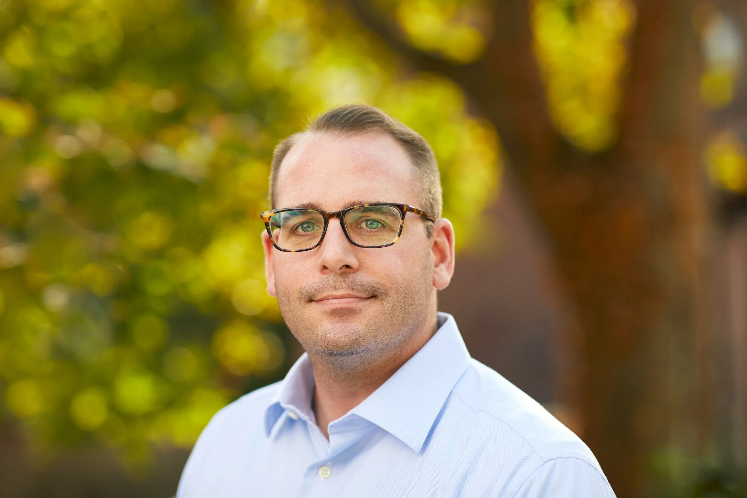 A man with short brown hair and glasses wearing a light blue button-up shirt standing outdoors with yellow and green blurred autumn leaves in the background.