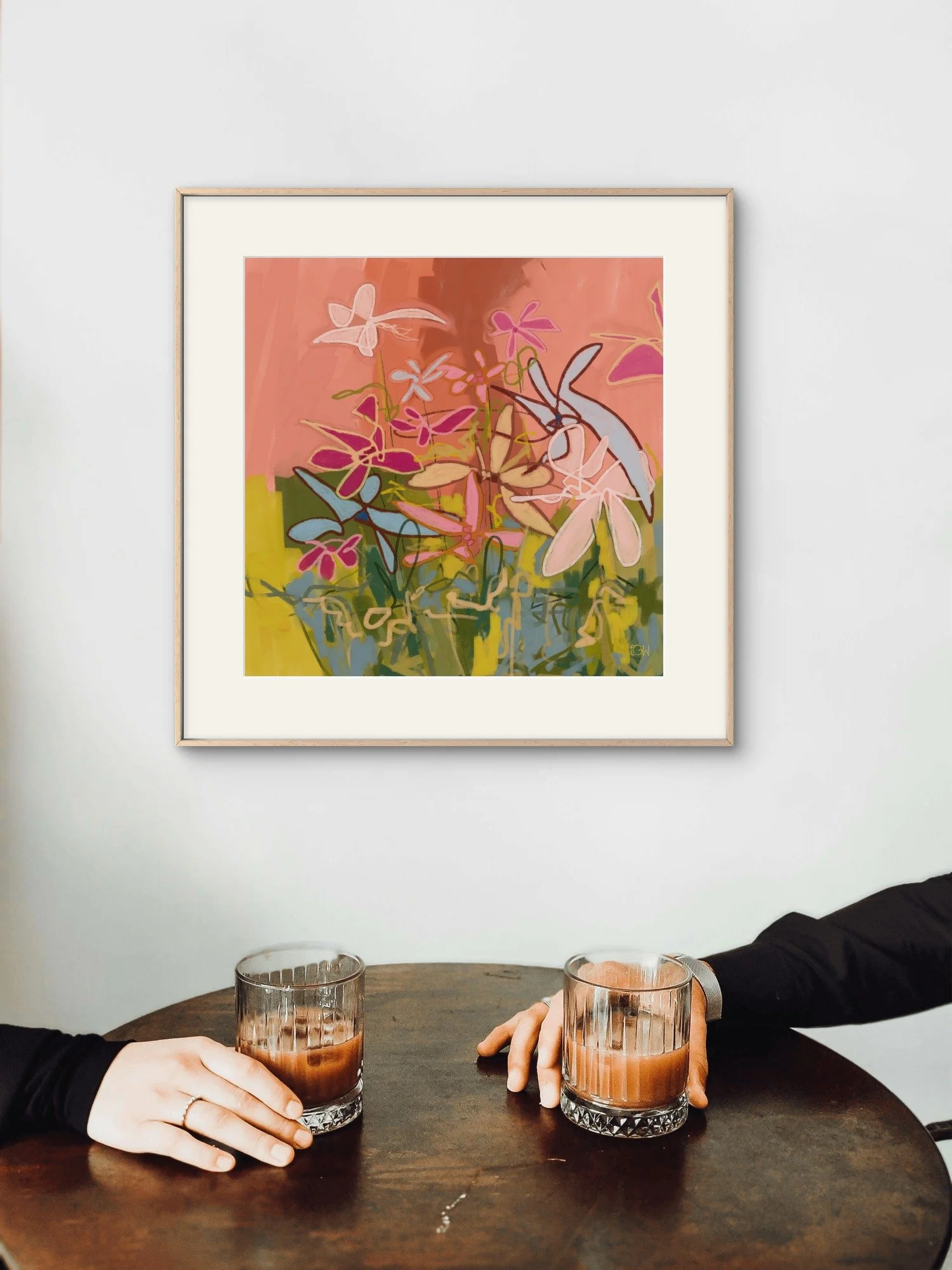 Two people, wearing black long-sleeve shirts, sit at a round table in a coffee shop toasting with coffee. In the background, a colorful abstract painting of flowers by Emily Whitish hangs on a white wall.