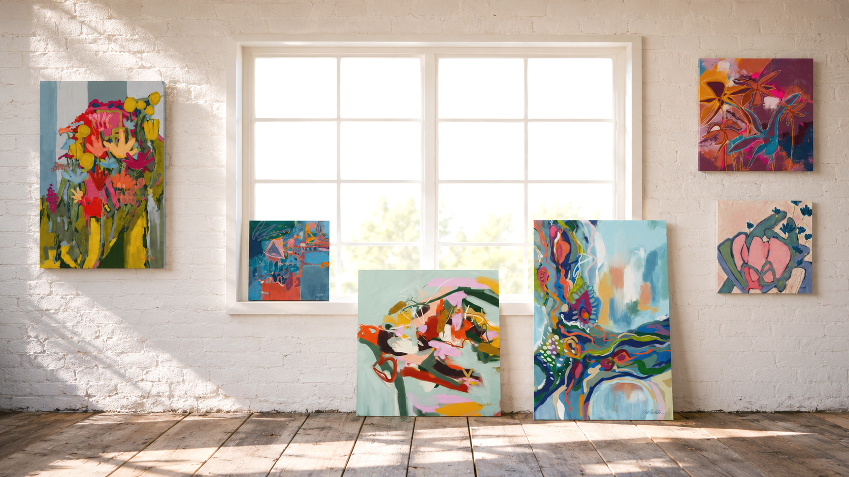 Studio scene featuring a collection of abstract paintings displayed on a white brick gallery wall with a large window, sunlight casting shadows on a wooden floor.